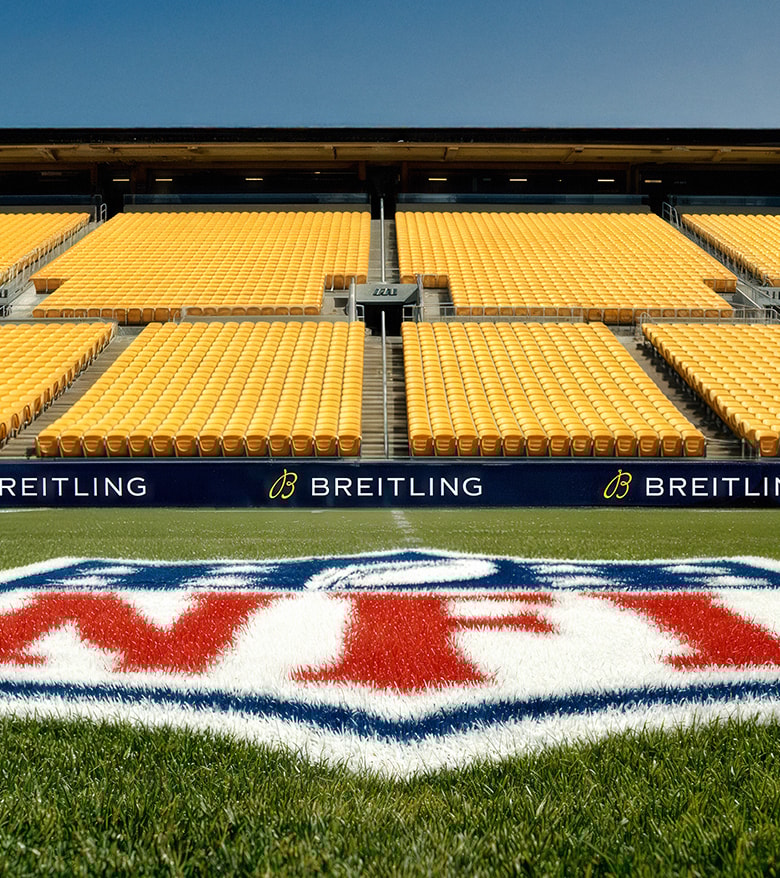 {generated} NFL pitch with bold midfield logo framed by empty yellow stands and clear Breitling branding along the touchlines.