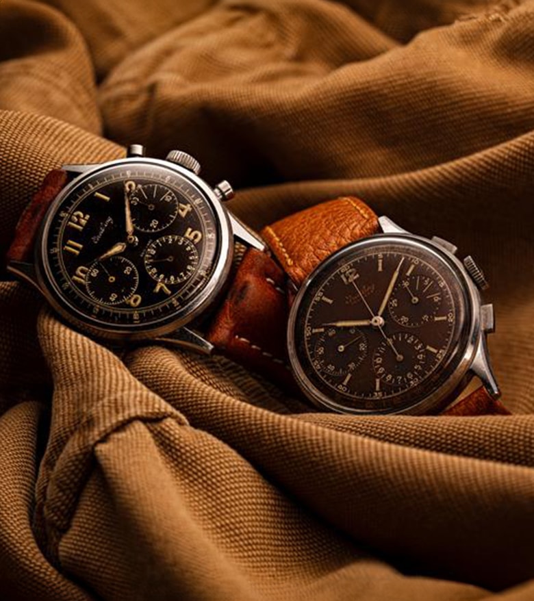 {generated} Two vintage Breitling Premier chronographs with dark dials and leather straps displayed against textured brown fabric.