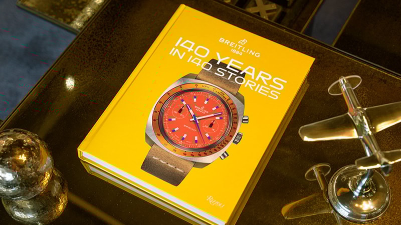 {generated} Yellow Breitling 140 Years book on a table, showing a red‑dial Breitling watch on its cover next to a small metal plane.