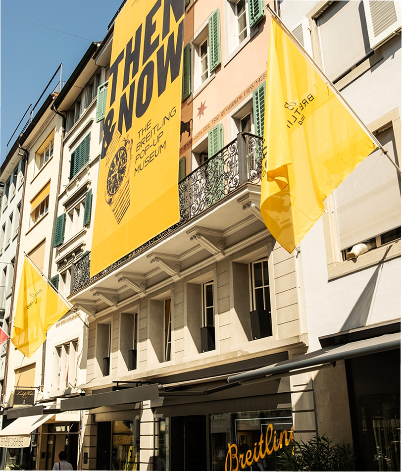 {generated} Facciata storica con vivide bandiere gialle Breitling che promuovono il museo modern‑retro sotto una luce soleggiata.