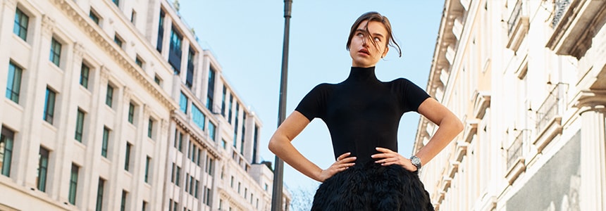 {generated} A woman poses in a black dress, showcasing a Breitling Lady Premier watch against an elegant city backdrop