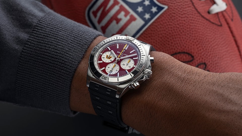 {generated} Breitling Chronomat com mostrador bordô Washington Commanders no pulso ao lado de uma bola oficial da NFL