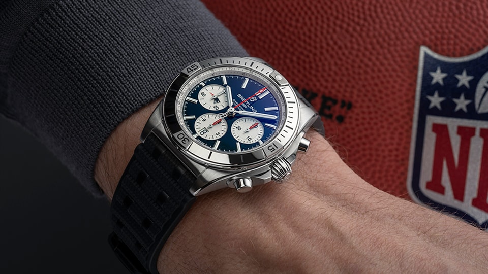 {generated} Cronógrafo Breitling com mostrador azul no pulso ao lado de uma bola de futebol, destacando o design dos New England Patriots.