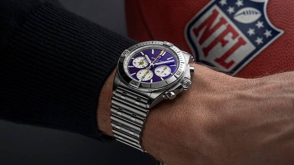 {generated} 佩戴的Breitling計時錶搭載明尼蘇達維京人紫色錶盤，置於NFL橄欖球旁，展現其團隊靈感設計。