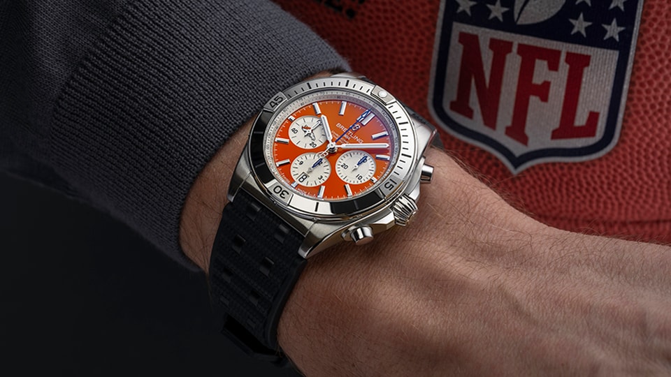 {generated} Breitling Chronomat with an orange dial on a dark strap worn near an NFL football, highlighting Denver team colors.