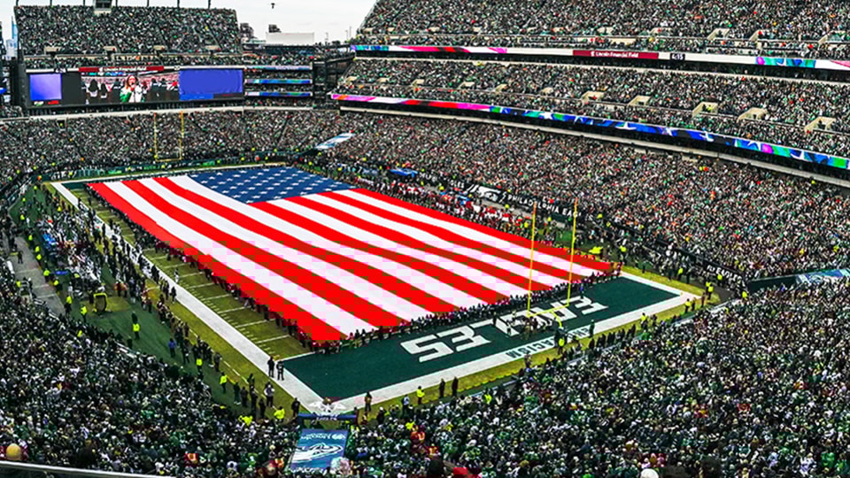 {generated} Un estadio repleto rodea una enorme bandera estadounidense que cubre el campo durante un partido de los Philadelphia Eagles.