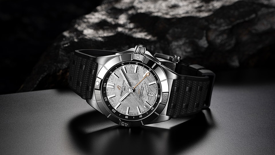 {generated} Breitling Chronomat Erling Haalandが質感あるシルバーダイヤルと黒いストラップで暗い岩肌の背景に映える。