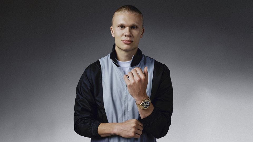 {generated} スポーティなジャケットの男性がゴールドのBreitling Chronomatを示し、その力強いデザインと精密さを際立たせている。