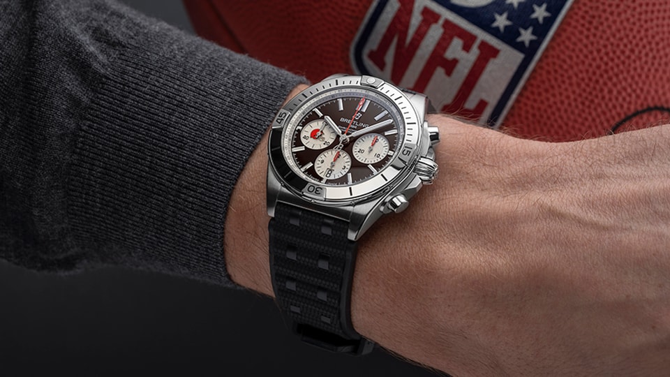 {generated} Хронограф Breitling на запястье с акцентами Cleveland Browns, размещён на фоне мяча NFL, подчёркивая спортивное партнёрство.