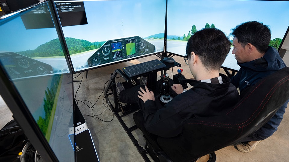 {generated} 二人の参加者がマルチ画面のフライトシミュレーターを操作し、計器類と滑走路表示に集中して訓練している様子。