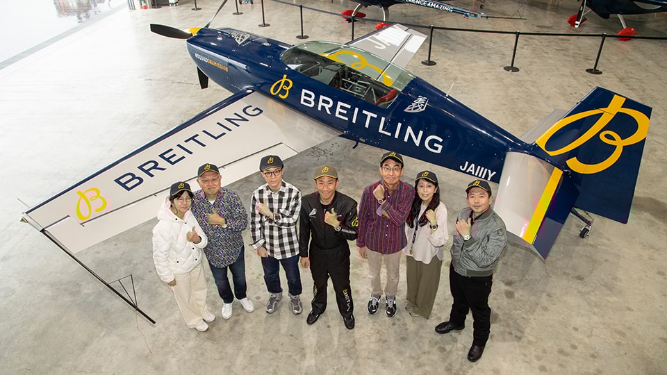 {generated} グループがBreitling仕様の曲技飛行機の前に立ち、広いハンガー内でのチャレンジフライトの様子を印象的に伝える構図