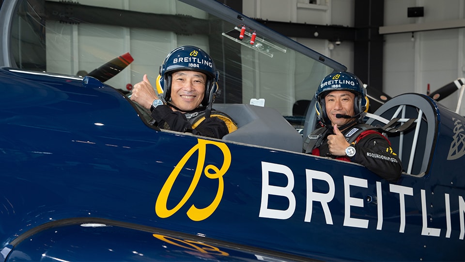 {generated} 2人のパイロットがBreitlingの曲技機で親指を立て、Breitlingヘルメットと腕元の時計を示している。