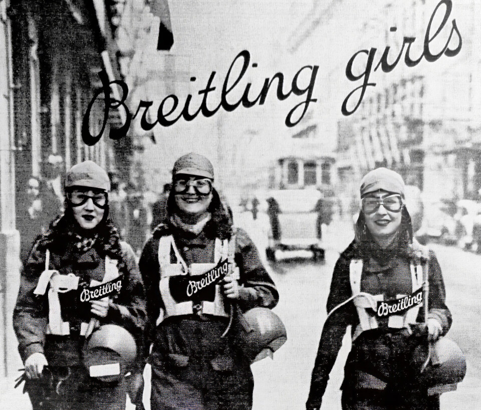 {generated} 三位身著復古航空裝備的女性走在城市街道上，醒目的Breitling標識展現品牌的傳統與風格