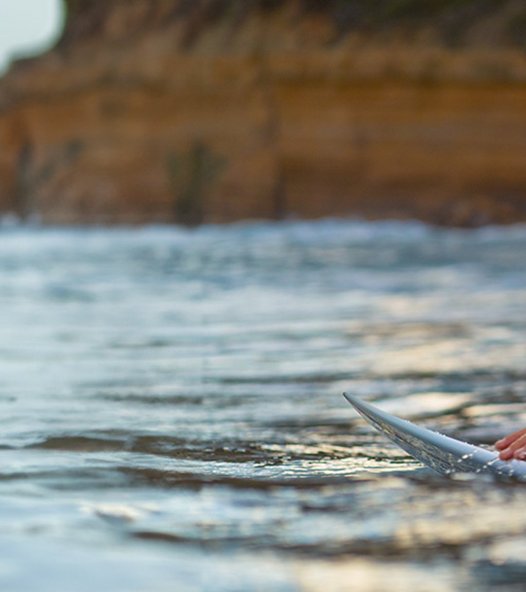 {generated} A woman paddling a surfboard at sunset, wearing a wetsuit and a Breitling watch, with coastal cliffs in the background.