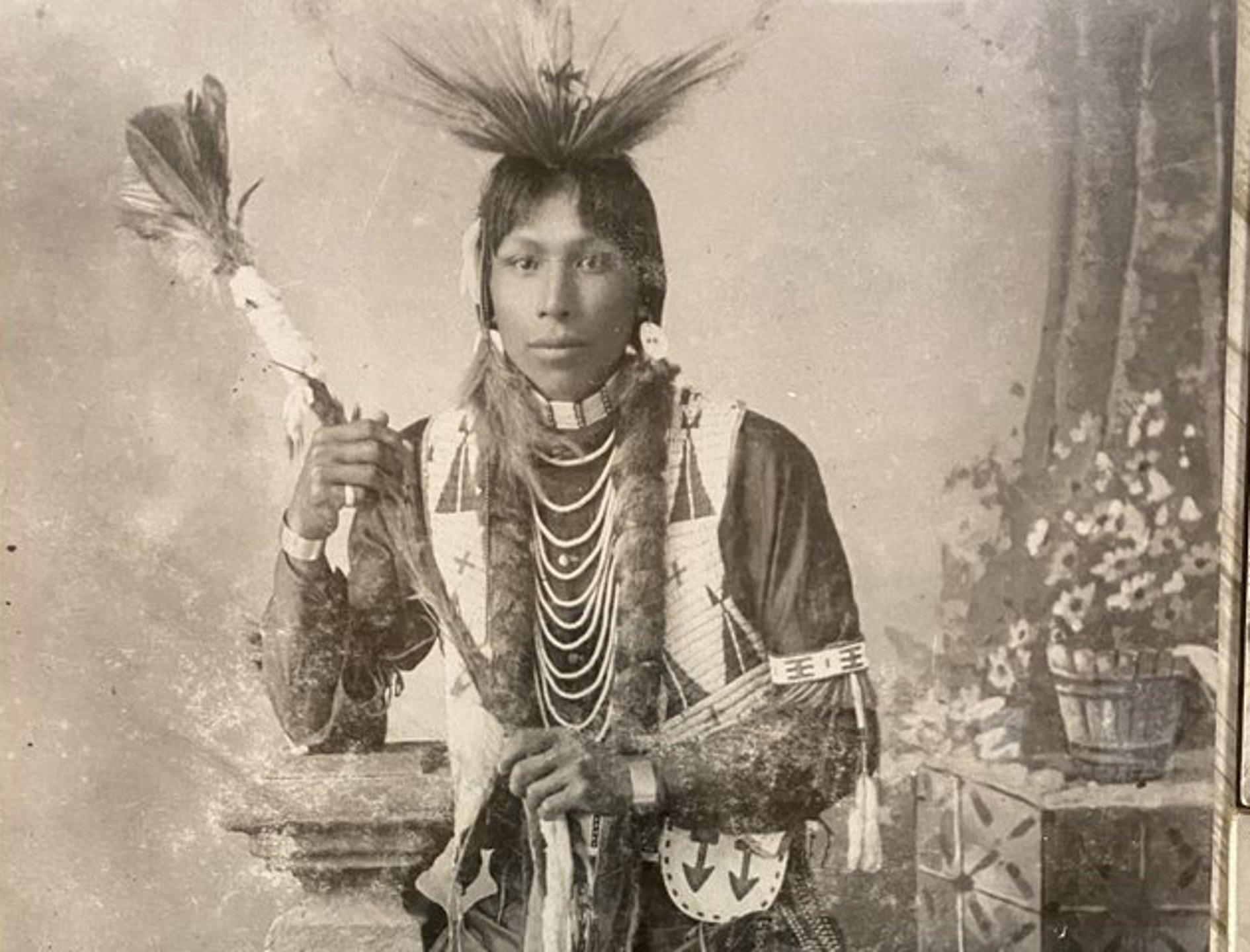 {generated} TU’PAXIN身穿傳統服飾並持裝飾手杖，站在具文化細節的攝影背景前。