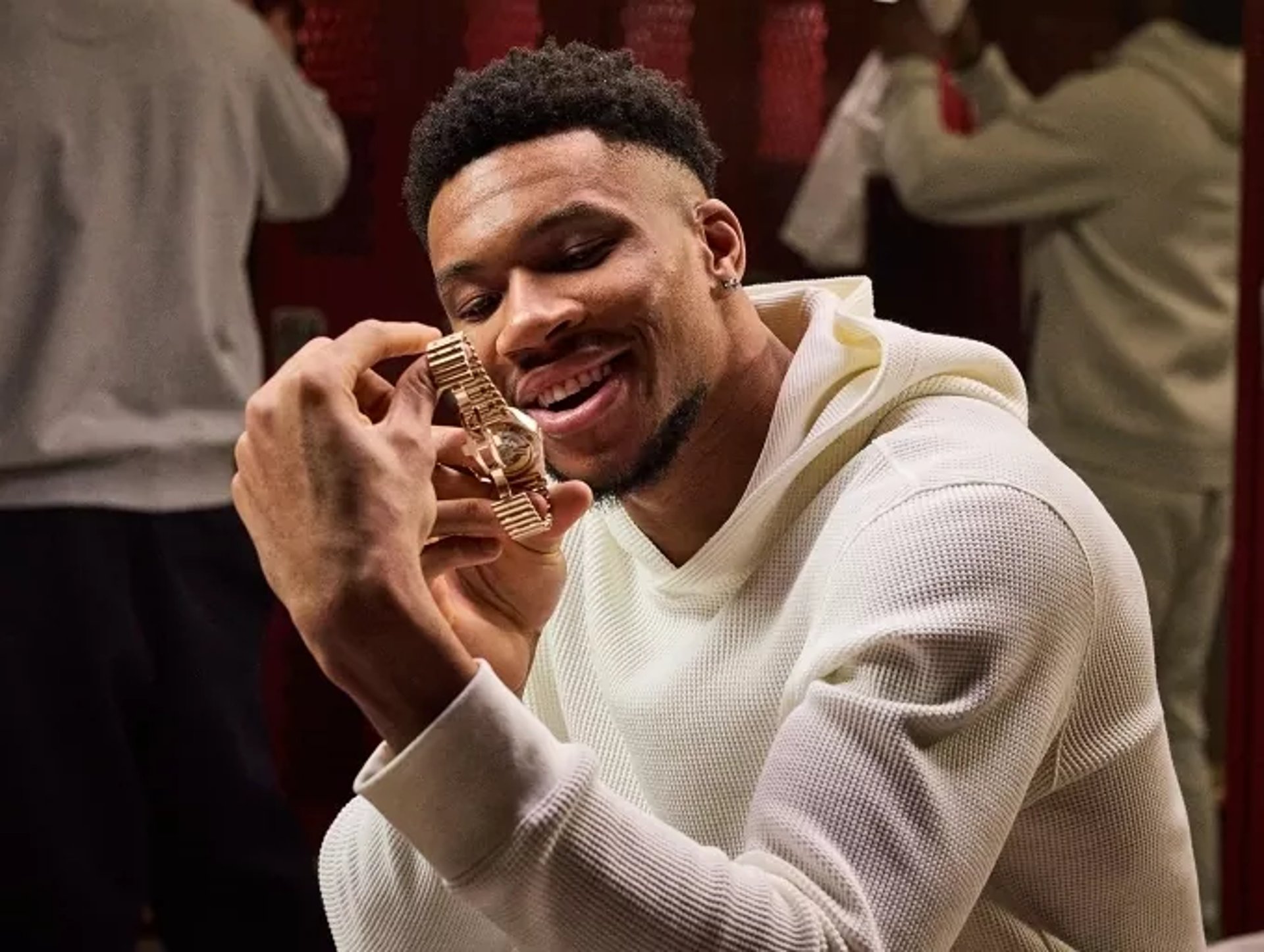 {generated} Giannis in a locker room adjusting his hoodie sleeve, shown in a warm athletic environment framed by reflective red lockers.
