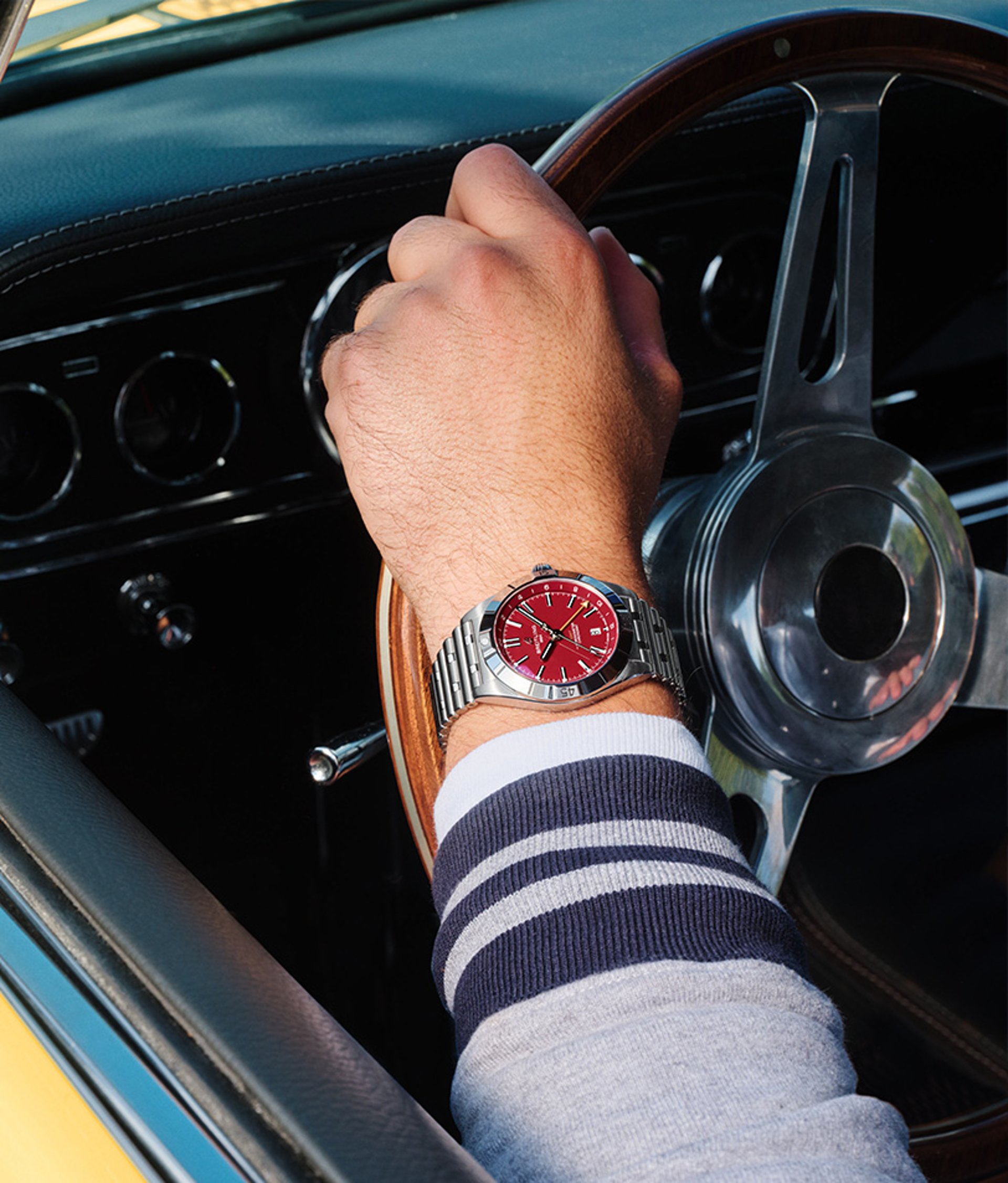 {generated} Driver's hand on a vintage steering wheel highlighting a Breitling Chronomat with a red dial and steel bracelet