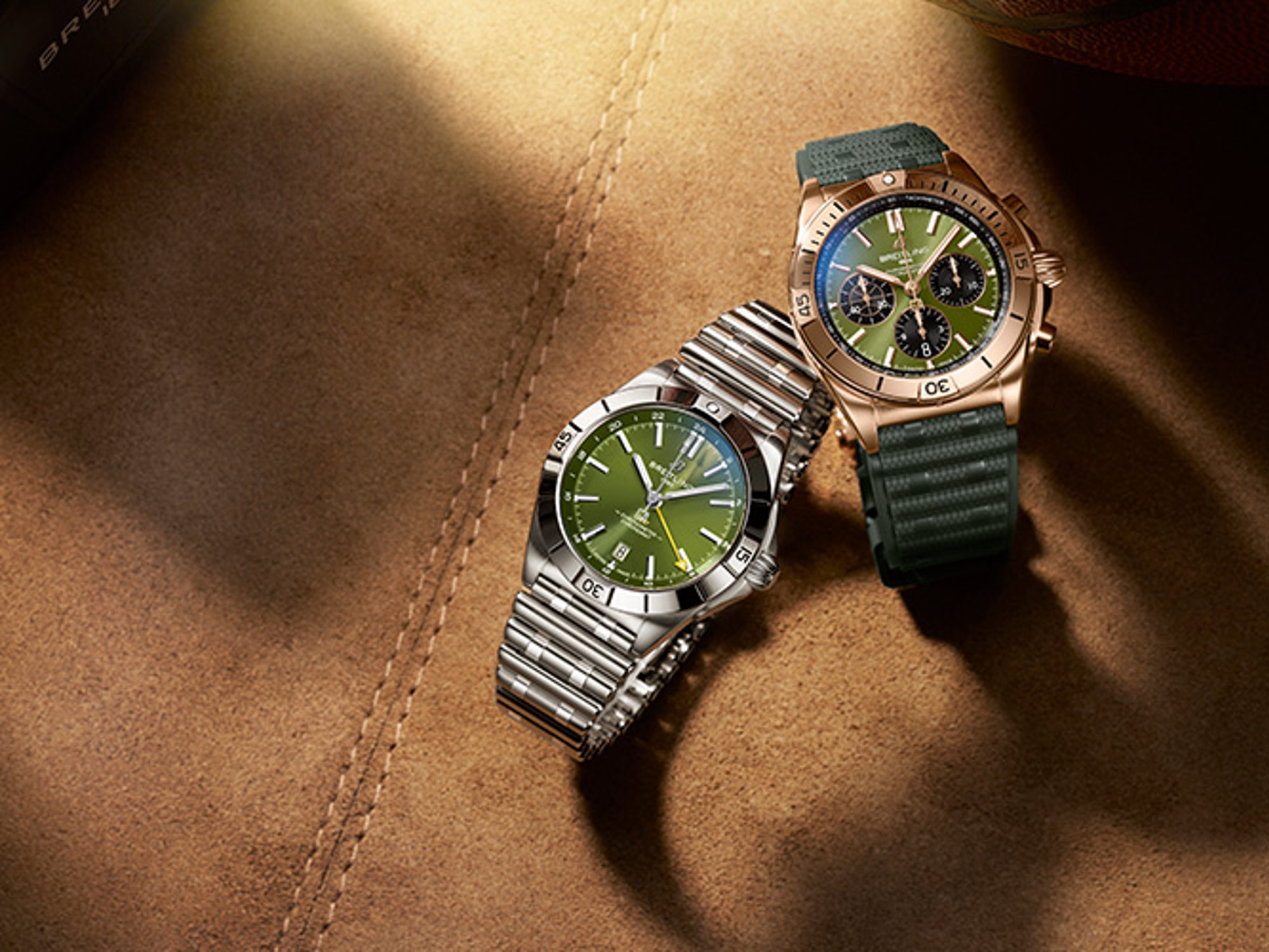 A gold and a silver Chronomat with dark green rubber straps on a brown table.