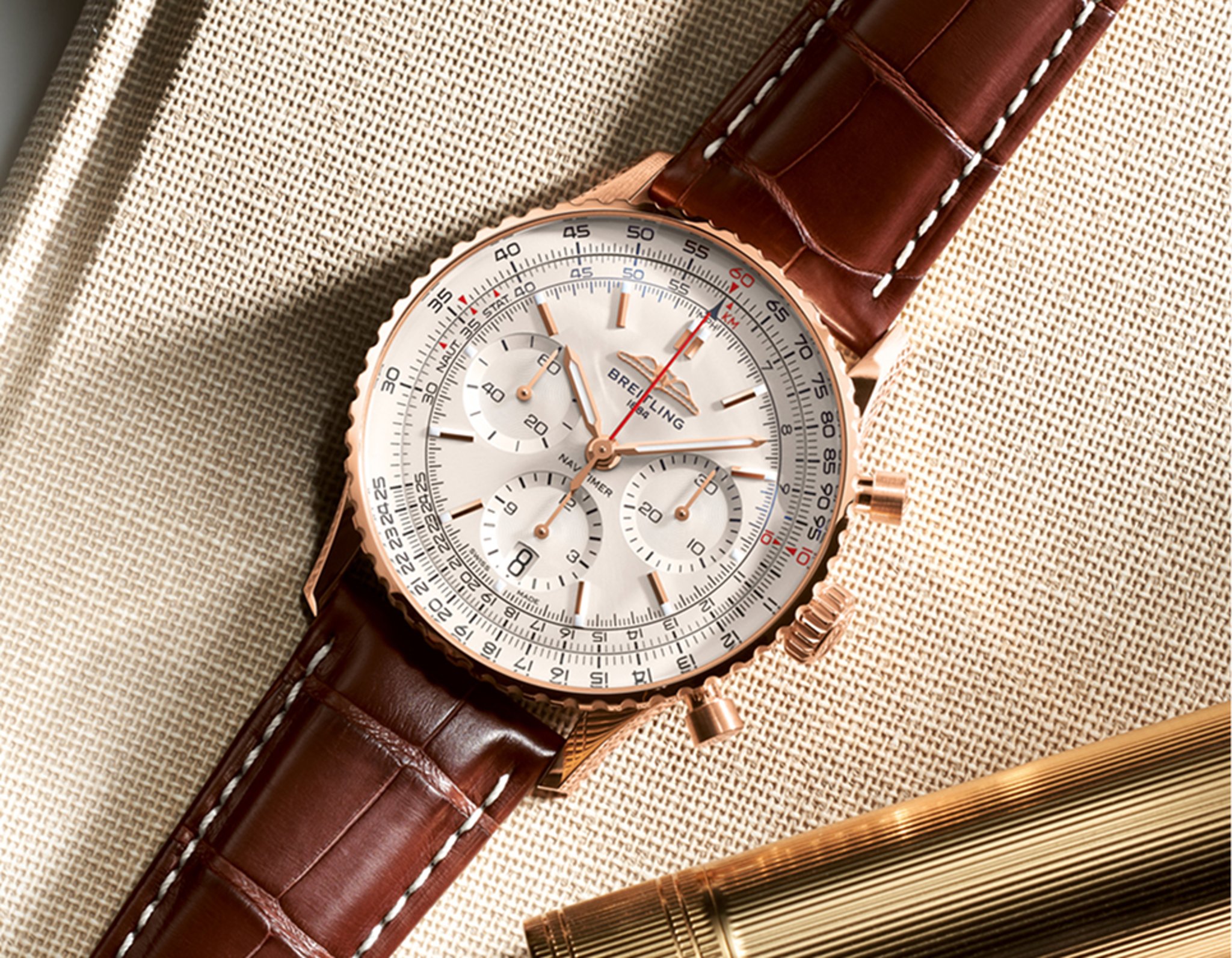 {generated} Breitling Navitimer in rose gold with a silver dial and brown leather strap displayed on textured fabric beside a gold pen.