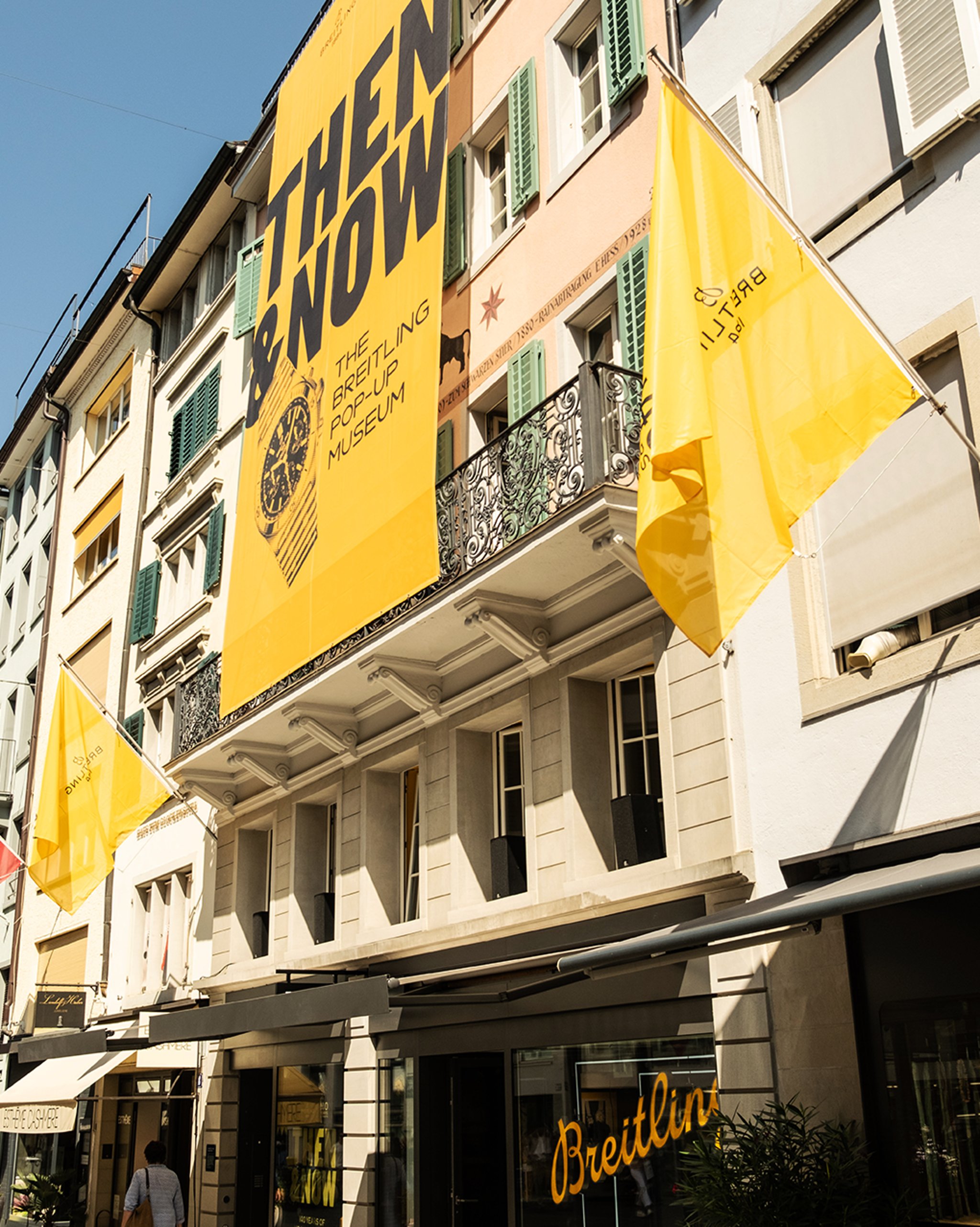 {generated} Street view of a Breitling storefront with bold yellow museum banners and flags highlighting the brand’s heritage.
