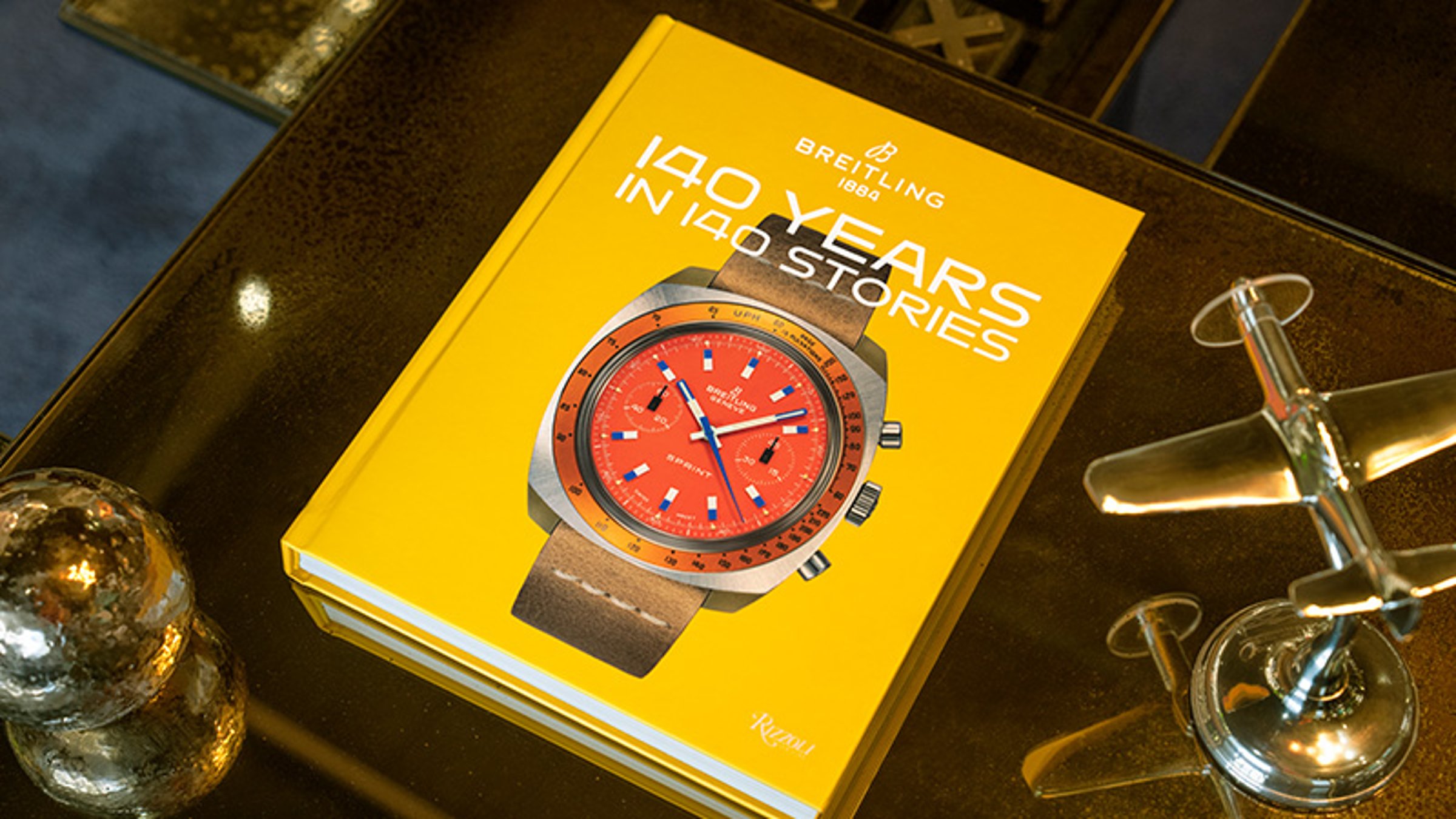 {generated} Yellow Breitling 140 Years book on a table, presenting a red‑dial Breitling watch on its cover beside a small metal plane.