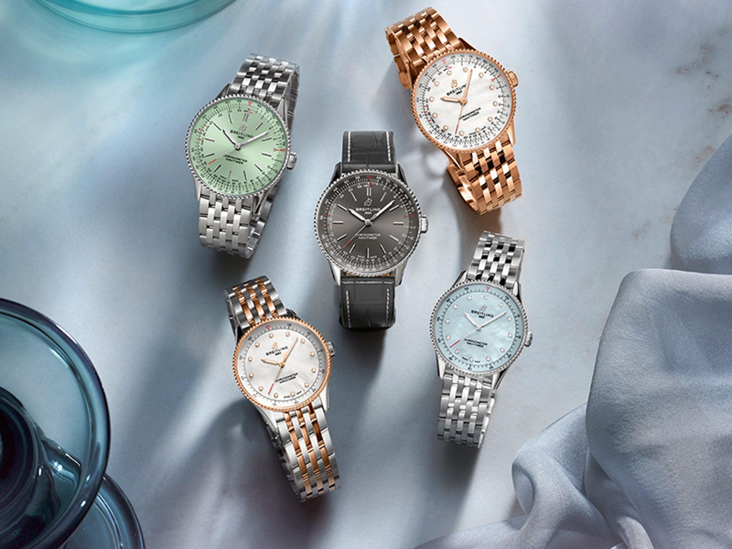 Five Navitimer watches with different-colored dials lie on a blue cloth.