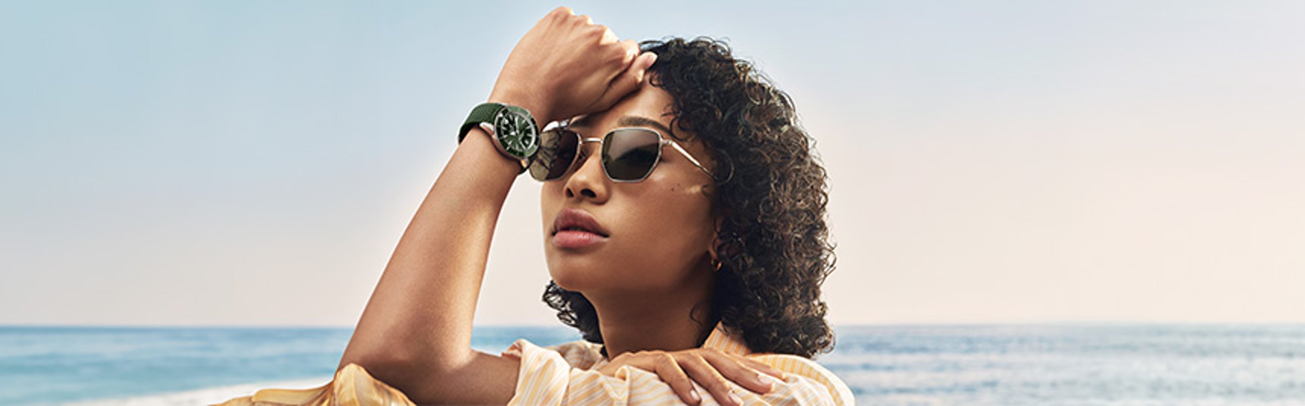 {generated} A person by the sea raises an arm, showcasing a green‑strap Breitling watch against a calm horizon in soft sunlight.