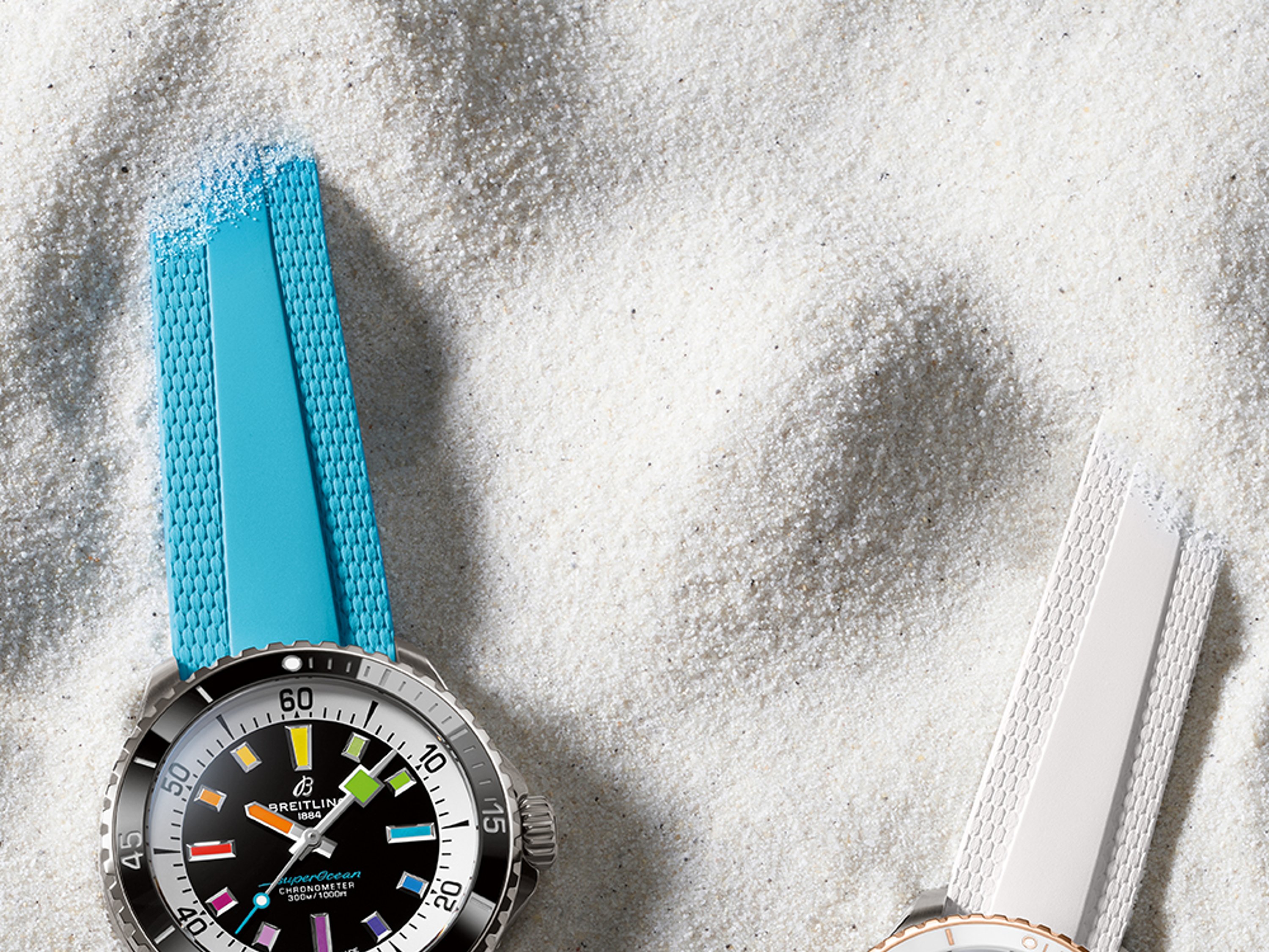 Two Superocean Automatic watches, one with a turquoise strap and one with a white strap, lie in the sand.