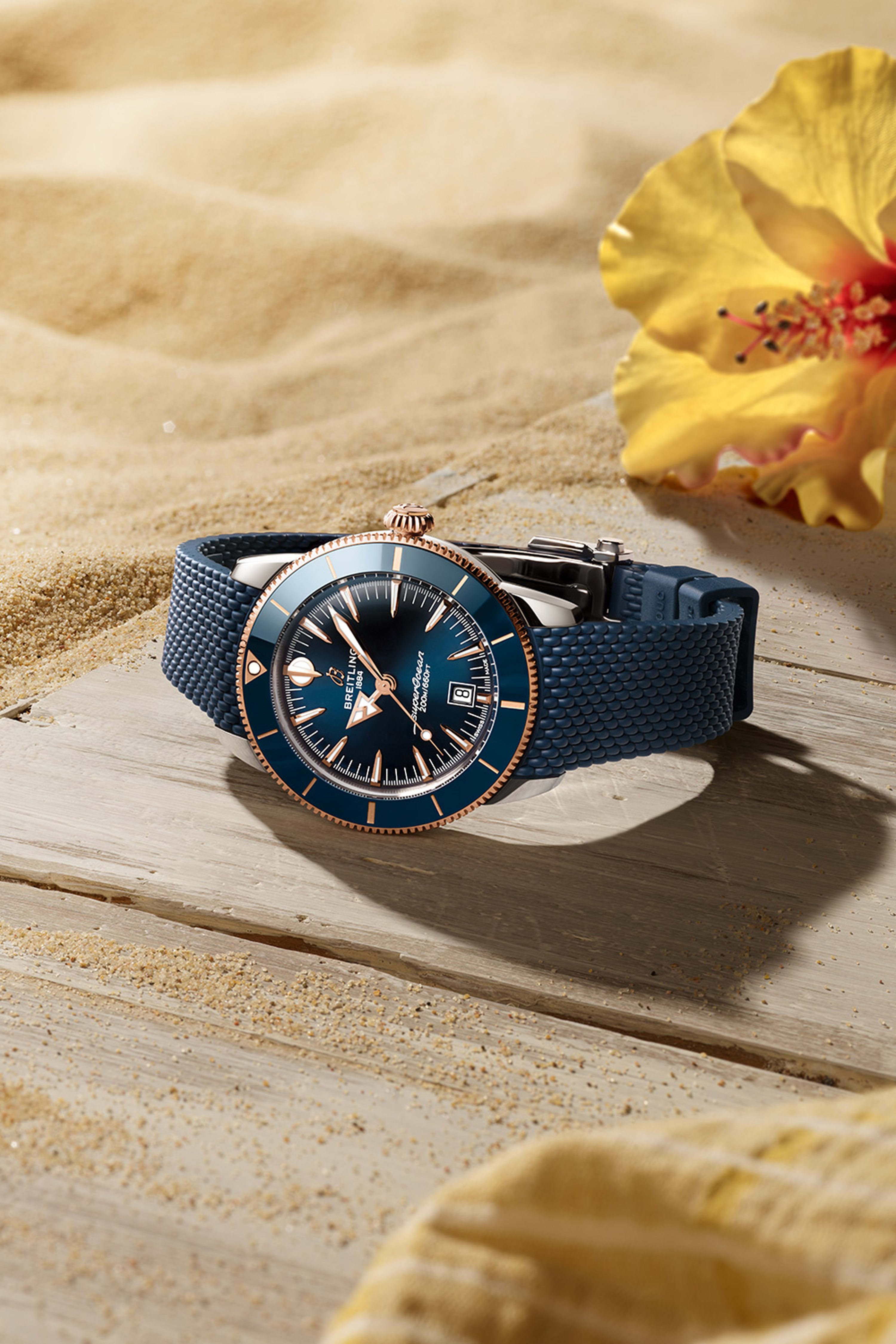 {generated} Breitling Superocean Heritage watch with blue dial and strap resting on sandy wood beside a bright yellow flower.