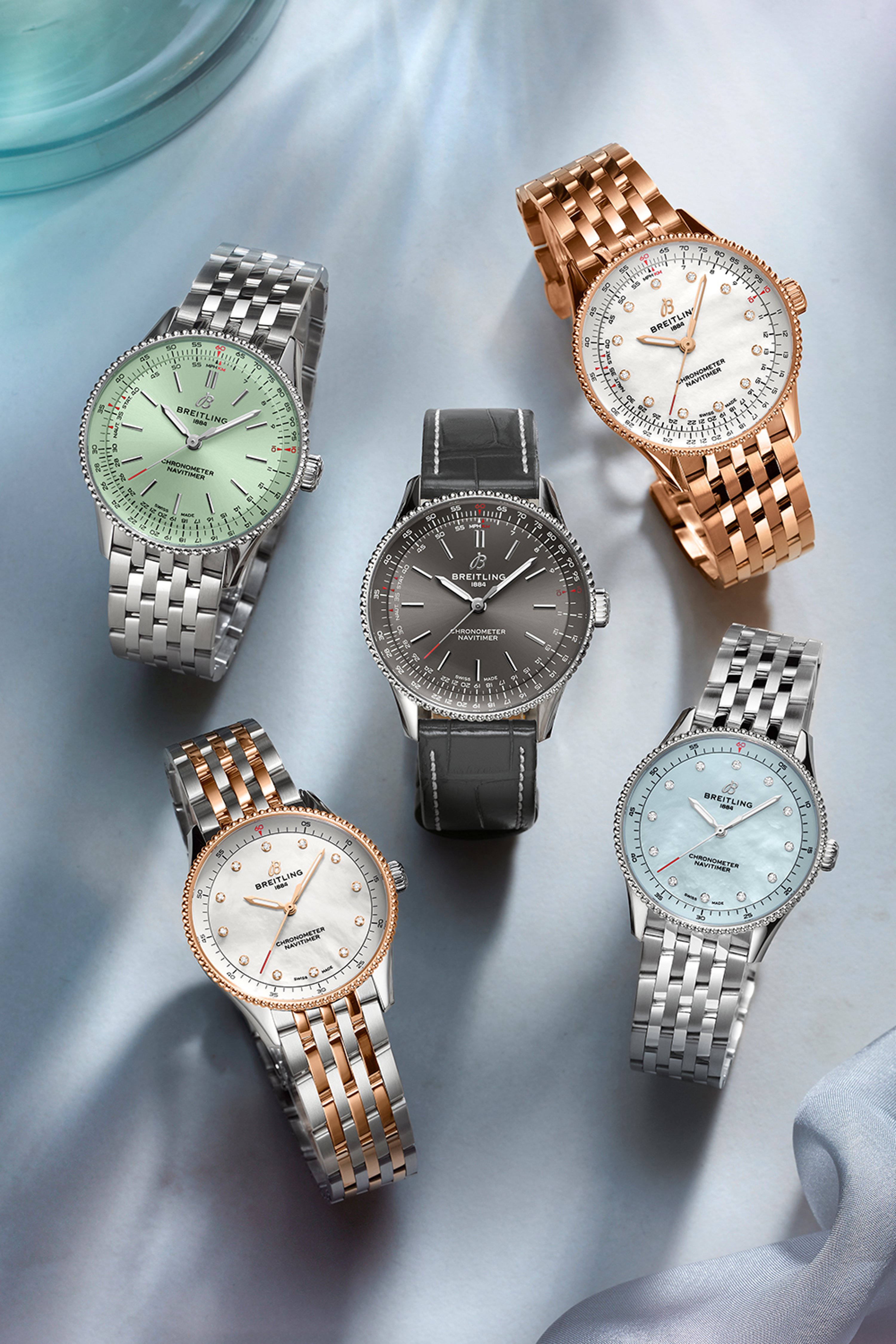 Five Navitimer watches with different-colored dials lie on a blue cloth.