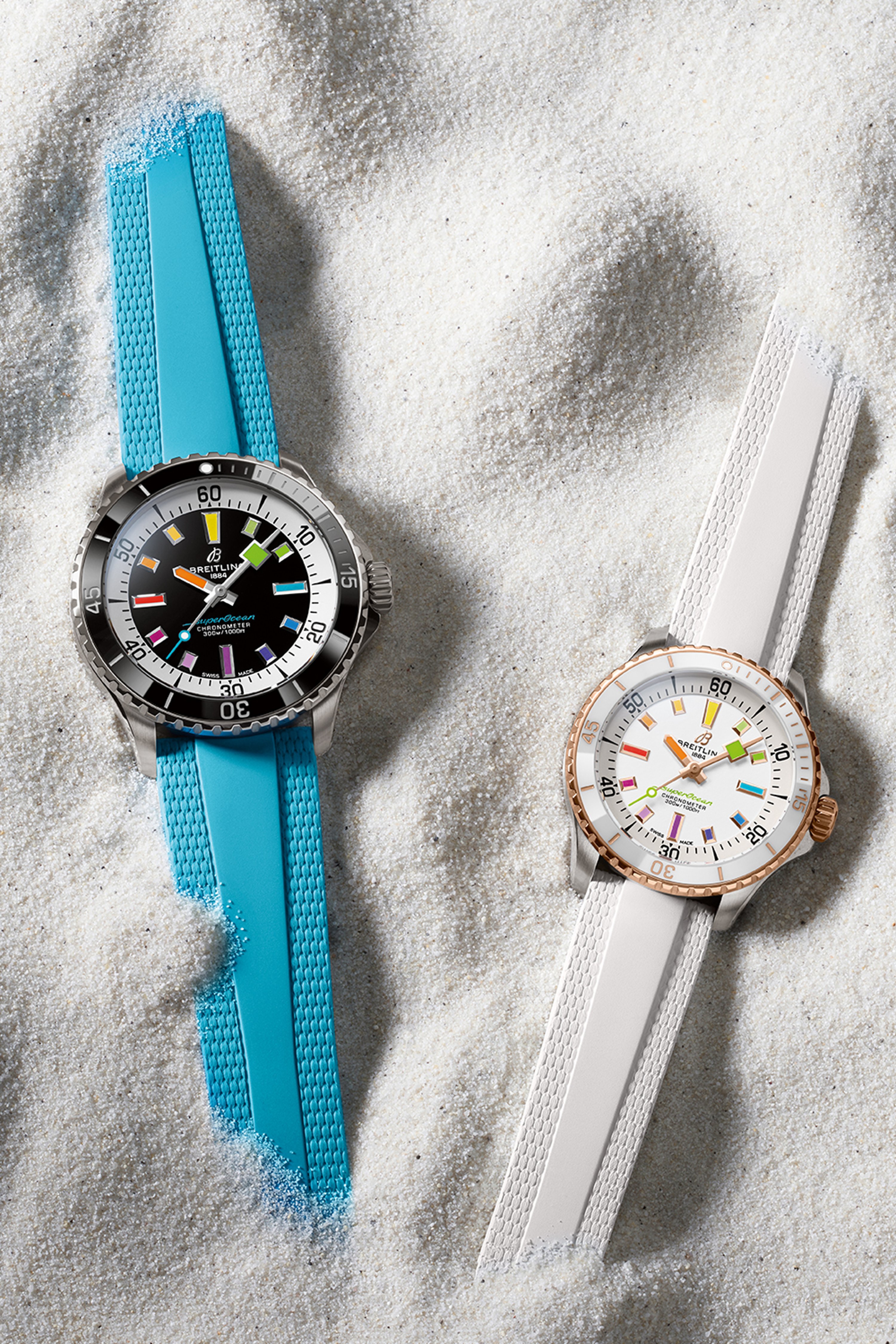 Two Superocean Automatic watches, one with a turquoise strap and one with a white strap, lie in the sand.