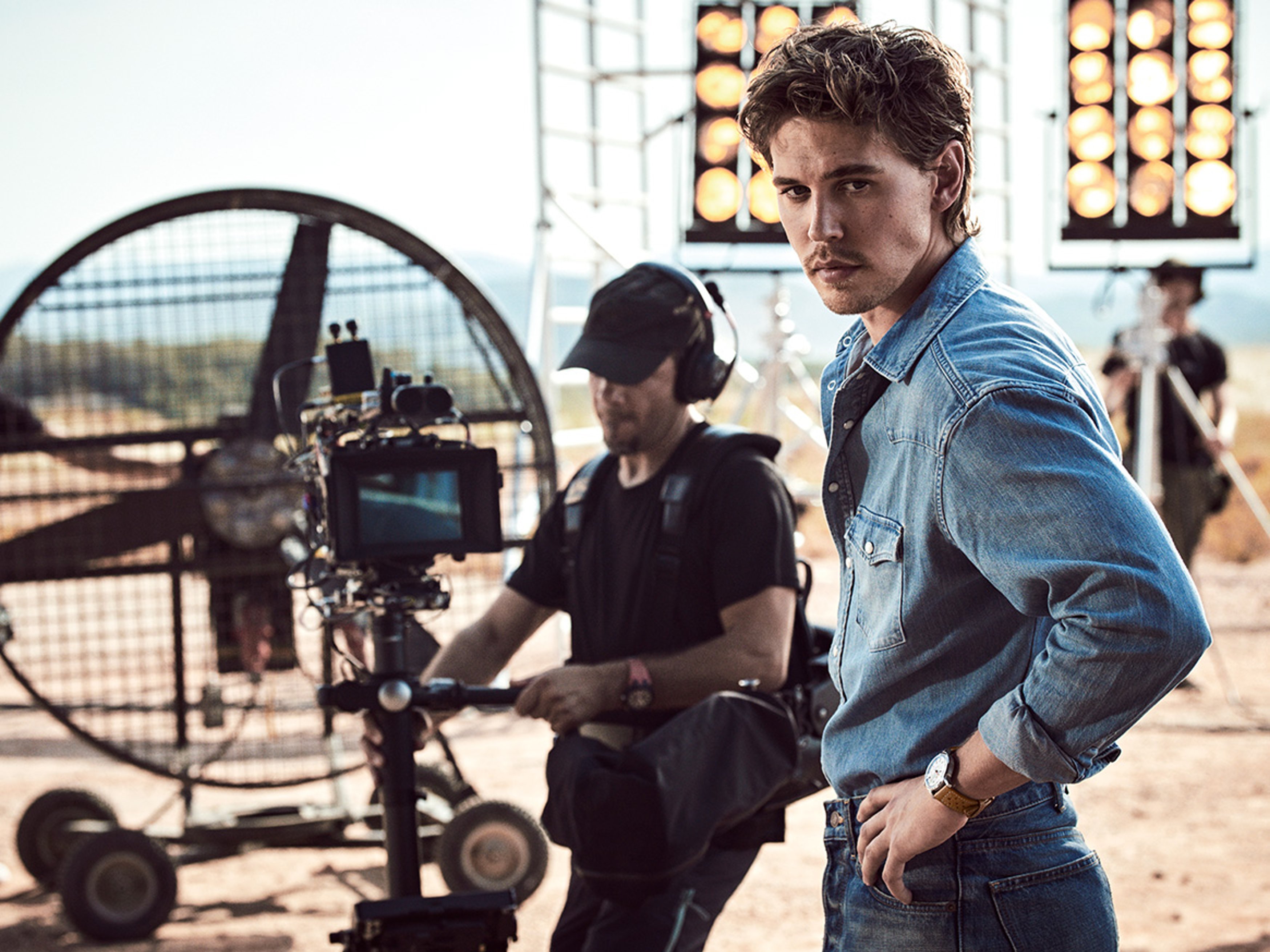 {generated} A male ambassador on a film set wearing a Breitling watch, standing beside crew and lighting equipment in a warm outdoor setting.