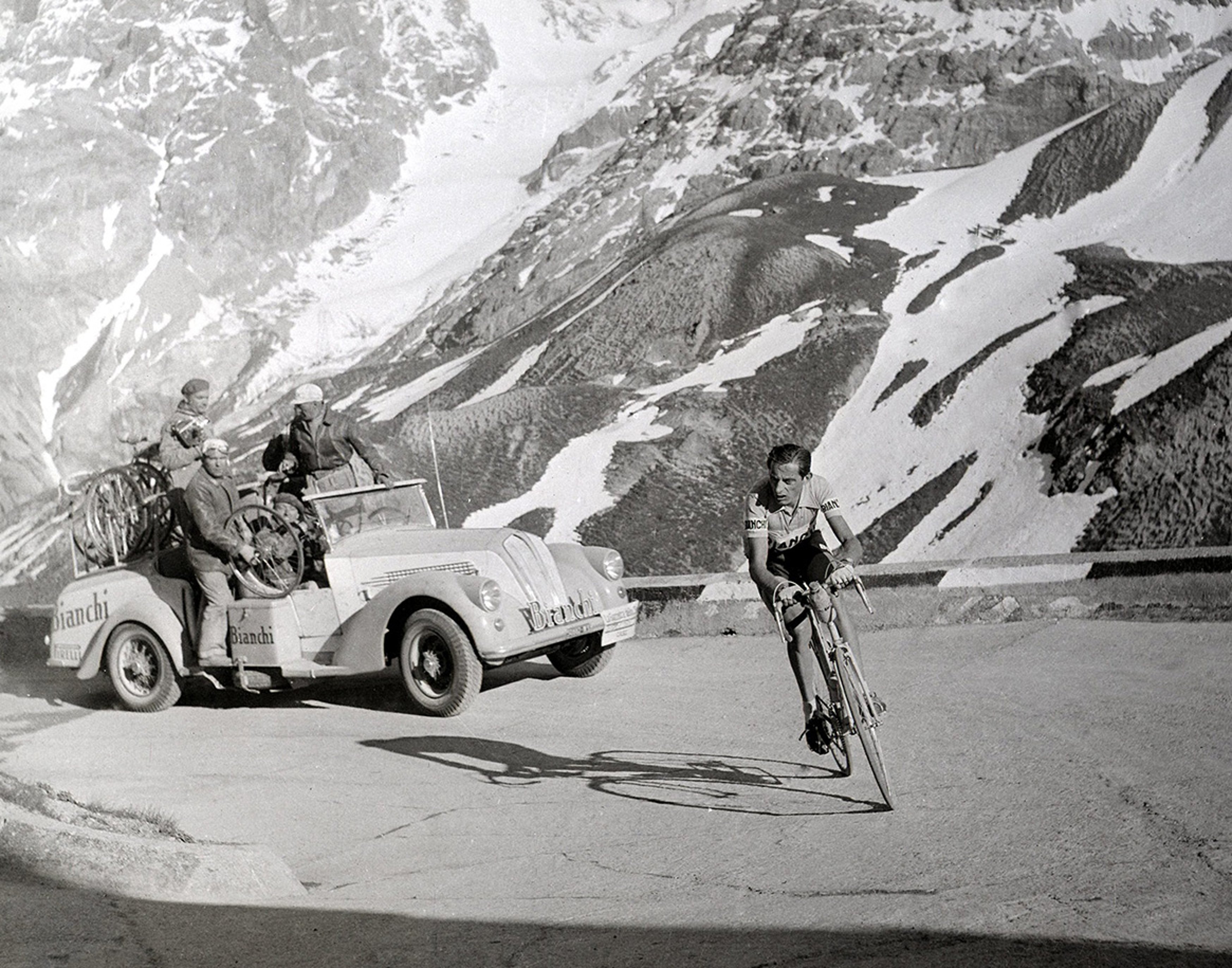 {generated} Vintage mountain road scene with a cyclist ahead of a support car by snowy peaks, capturing a historic race moment.