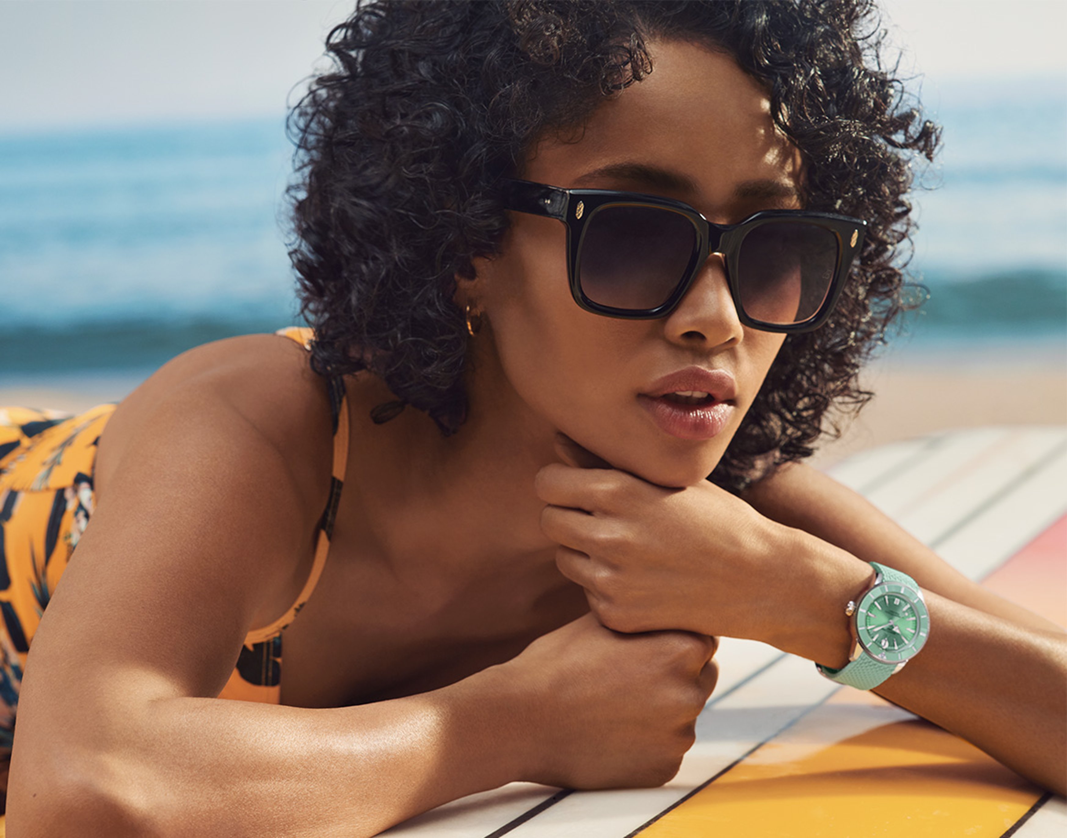 {generated} A woman relaxing on the beach wearing sunglasses and a turquoise Breitling Superocean watch resting on a surfboard.