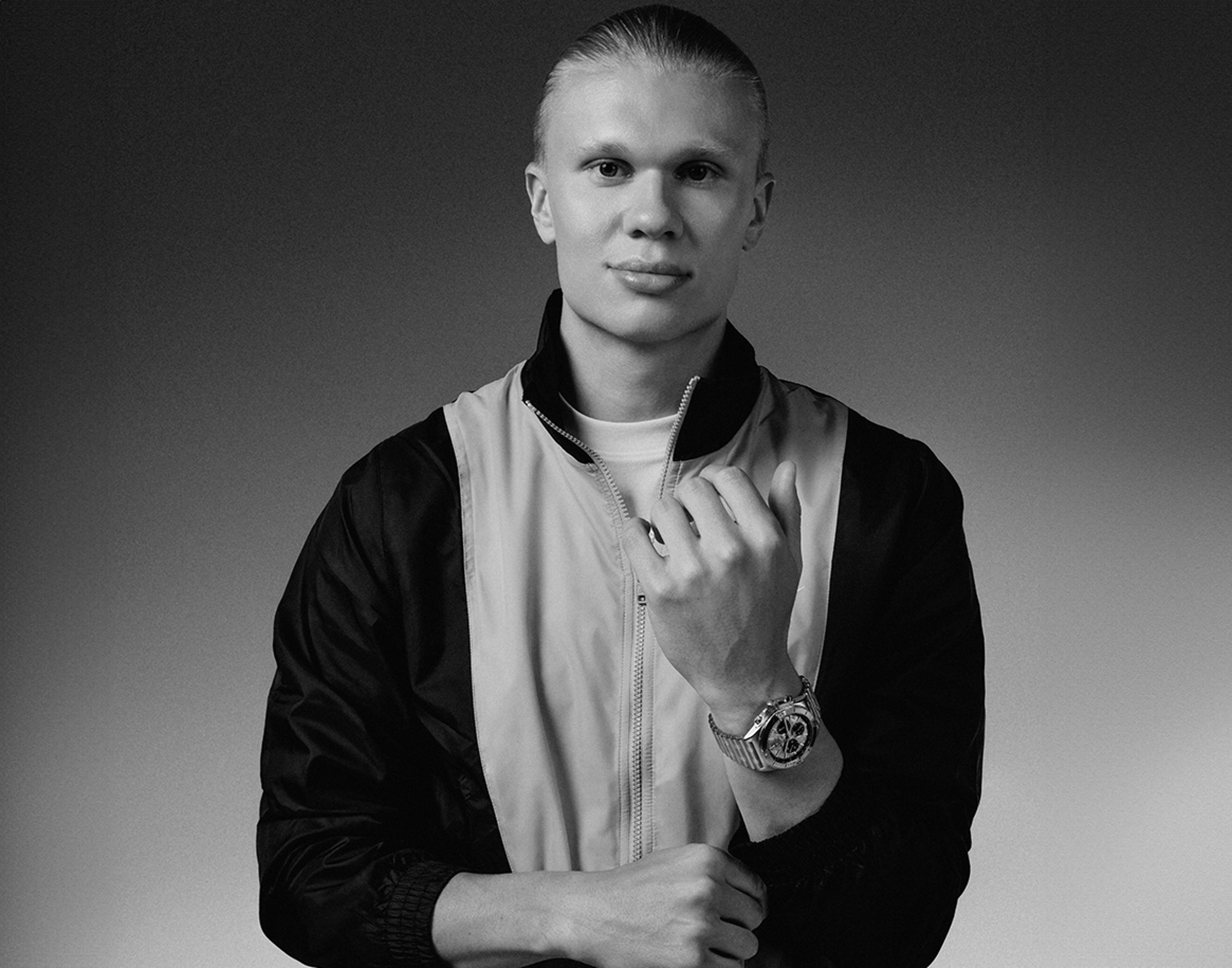 {generated} A man, noted in the title, wearing a sporty jacket and presenting a Breitling watch in a monochrome studio setting.