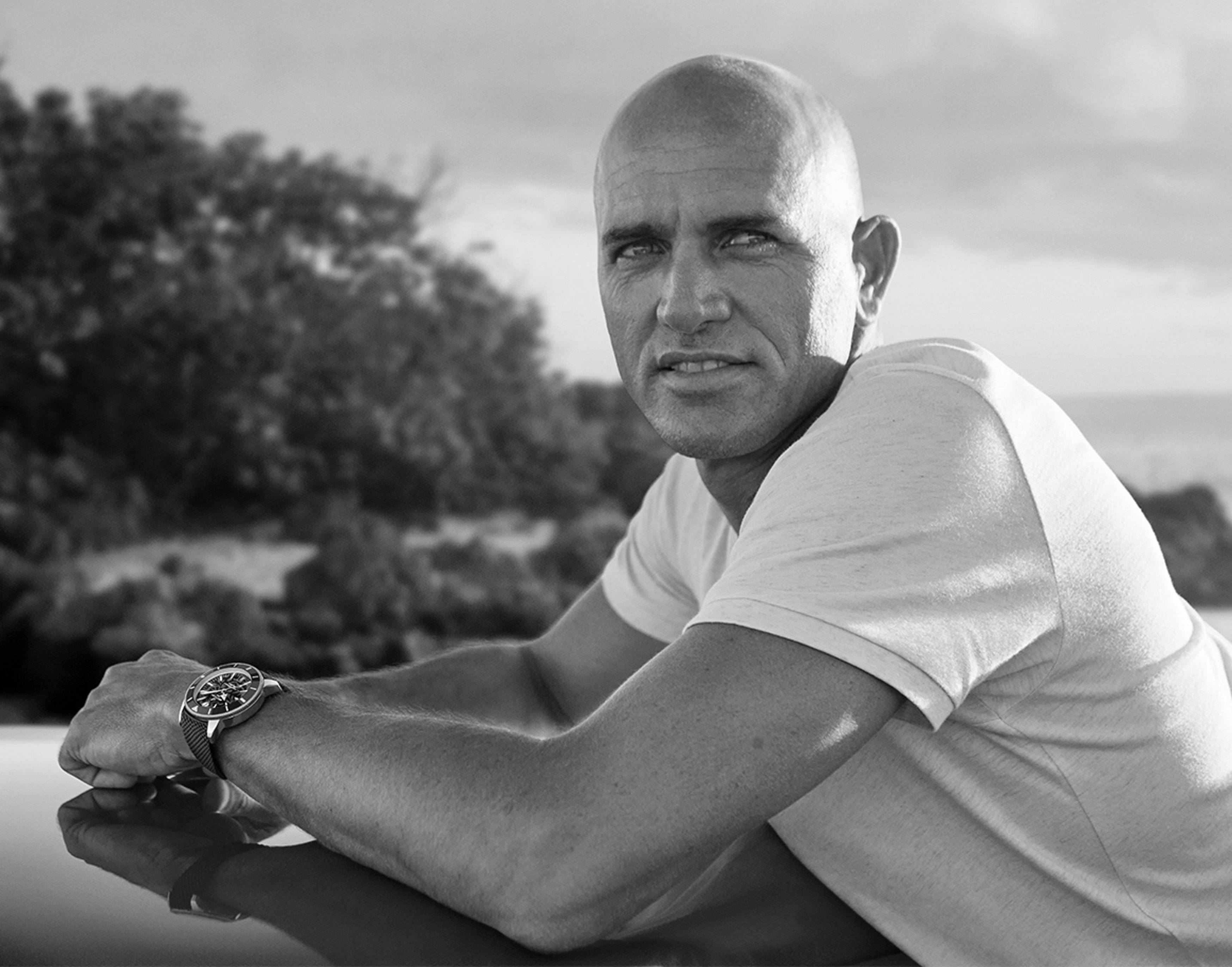 {generated} Kelly Slater outdoors wearing a Breitling watch, its bold dial visible as he rests his arms on a smooth reflective surface