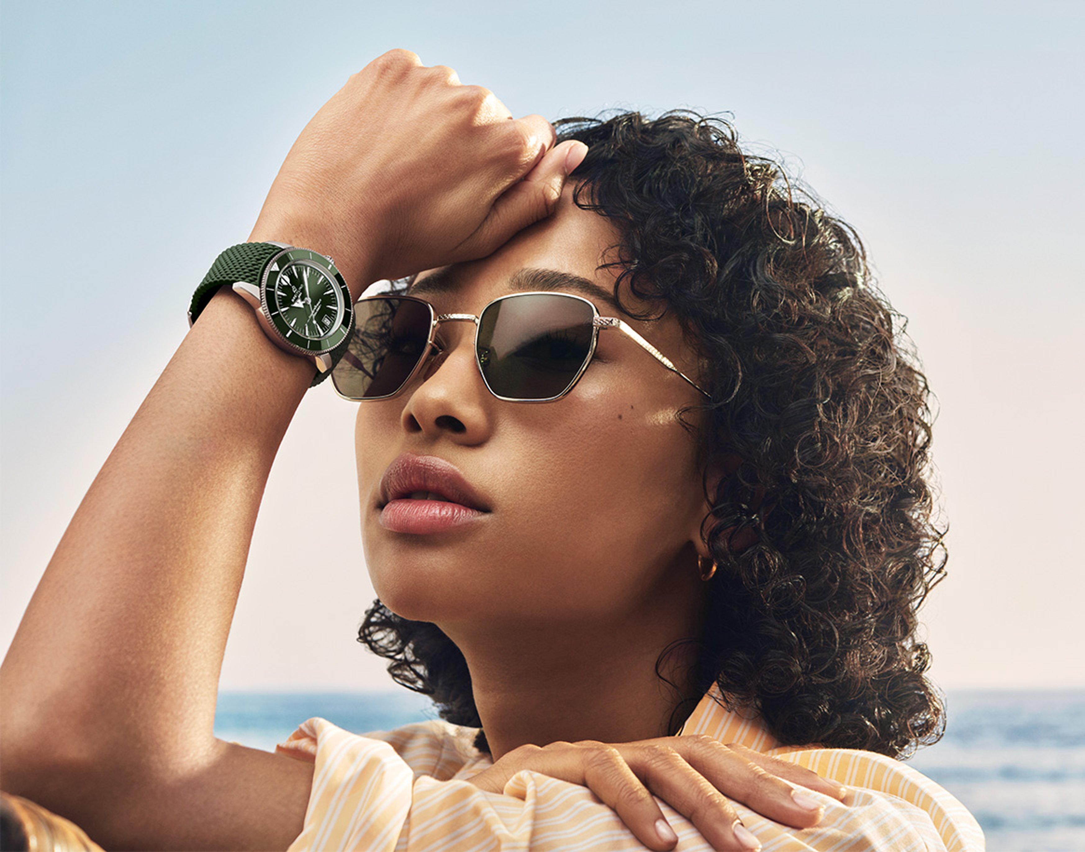 {generated} A woman by the sea lifts sunglasses as a green‑strap Breitling watch catches the light, highlighting relaxed summer style.