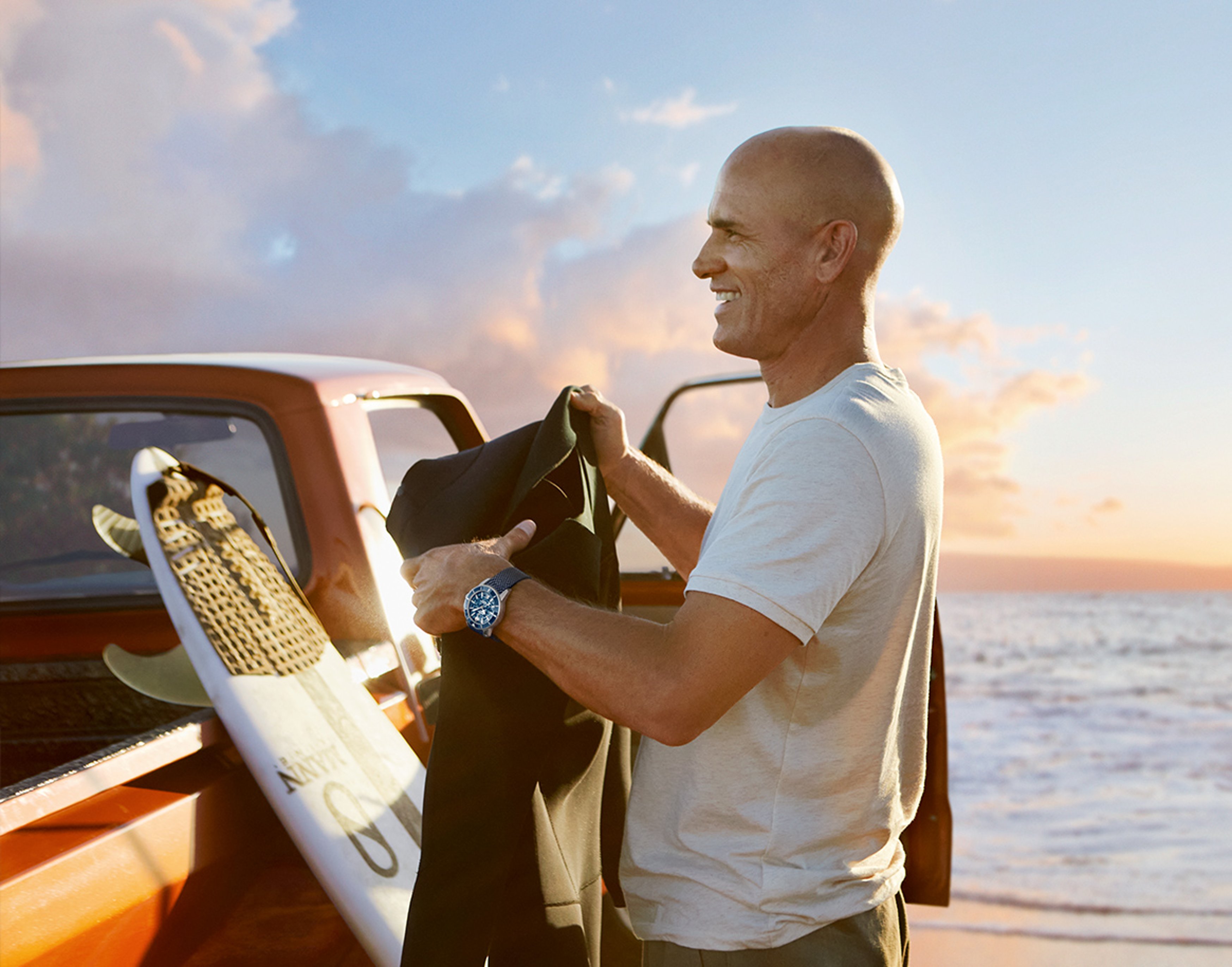 {generated} Man by a truck at sunset holding a wetsuit, wearing a Breitling watch beside a surfboard on the beach.