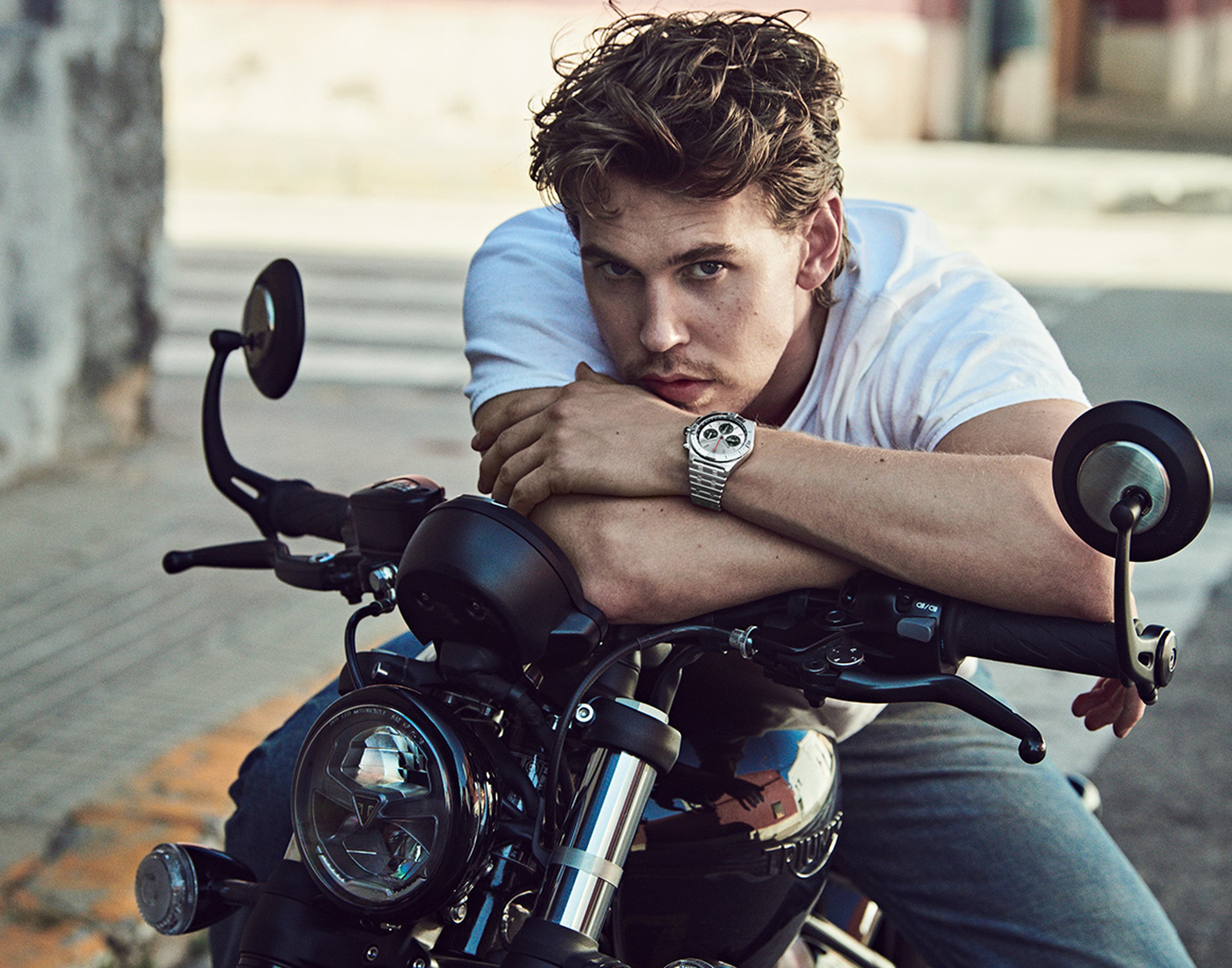 {generated} Austin leans on a motorcycle, displaying a Breitling Chronomat watch on his wrist in a relaxed outdoor urban setting.