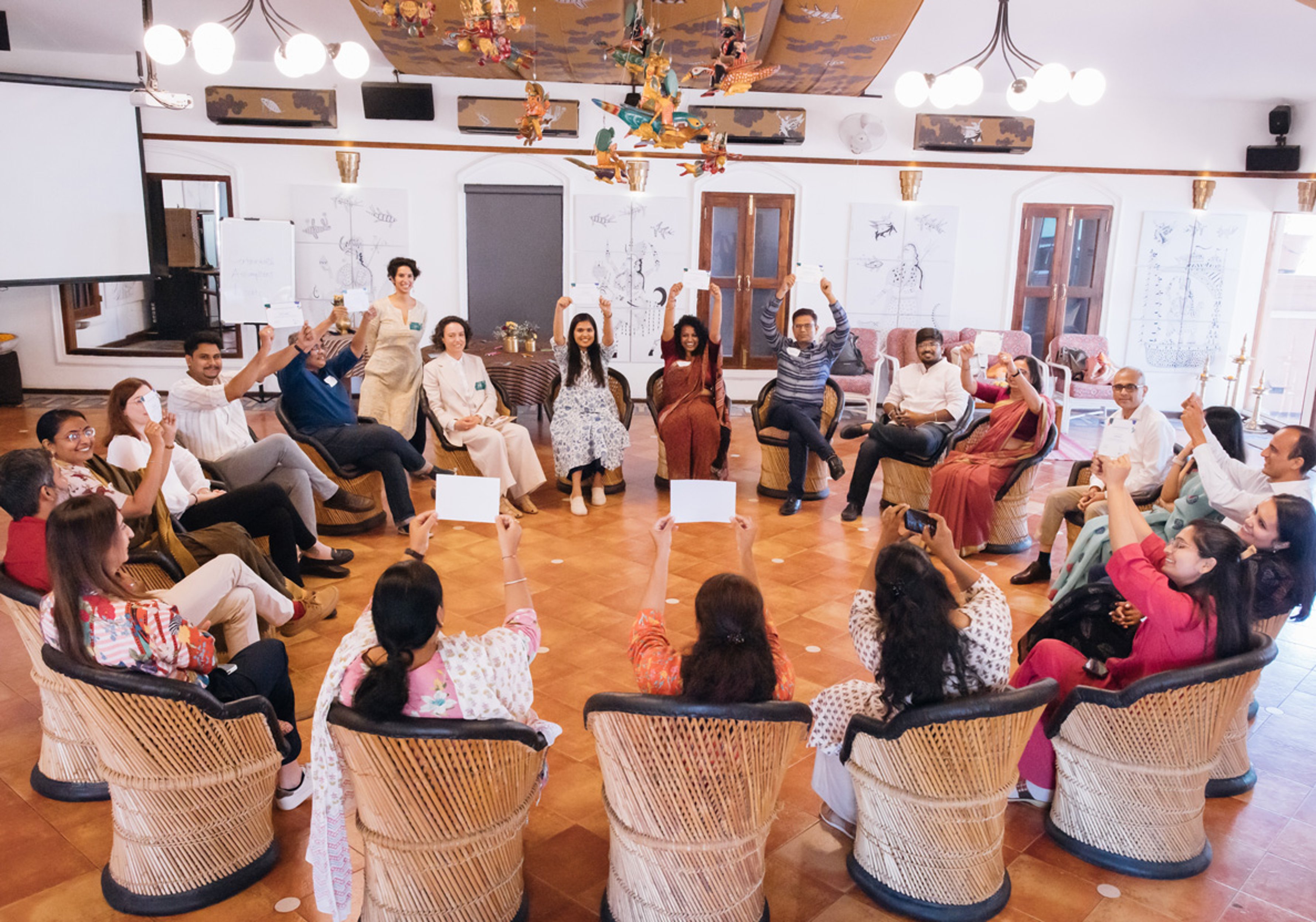 {generated} Un groupe divers assis en cercle lève des cartes blanches dans une salle lumineuse tout en participant à une activité communautaire.
