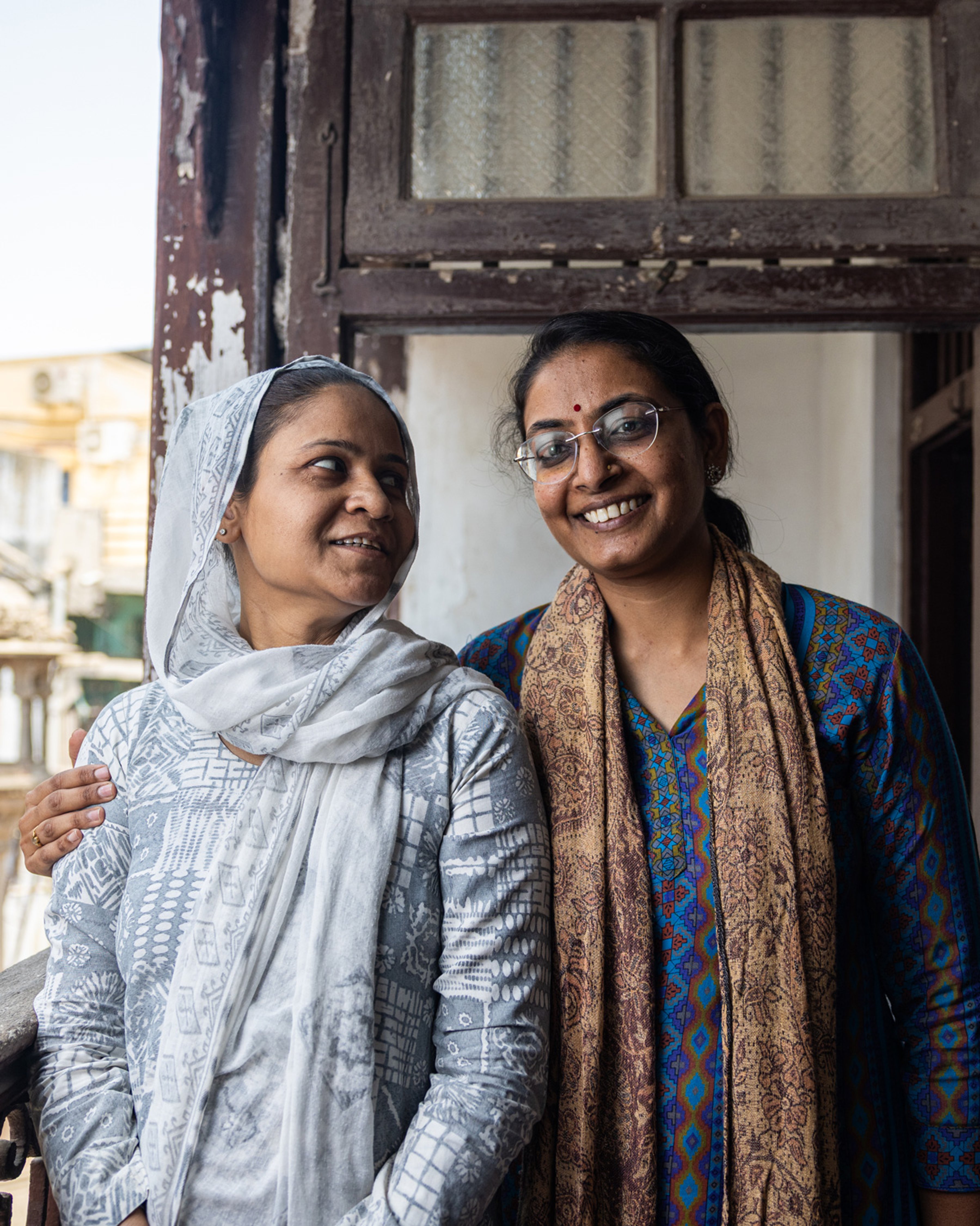 {generated} Deux femmes se tiennent côte à côte sur un balcon usé, leurs foulards ornés et leur attitude calme exprimant une solidarité mutuelle.