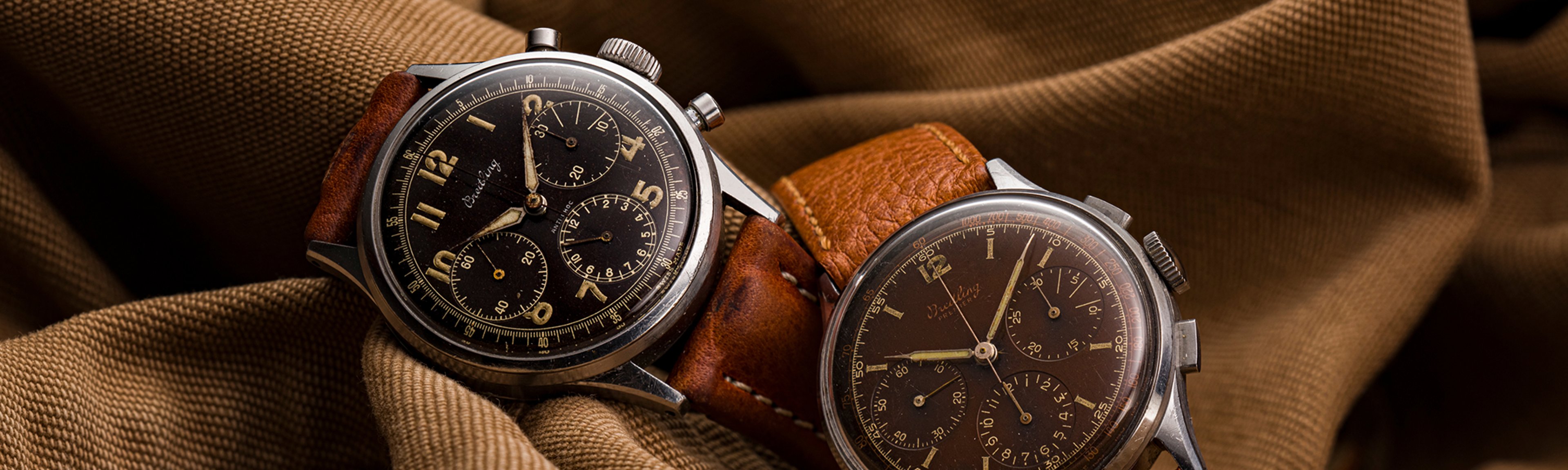 {generated} Two vintage Breitling Premier chronographs with dark dials and leather straps presented against softly textured brown fabric.