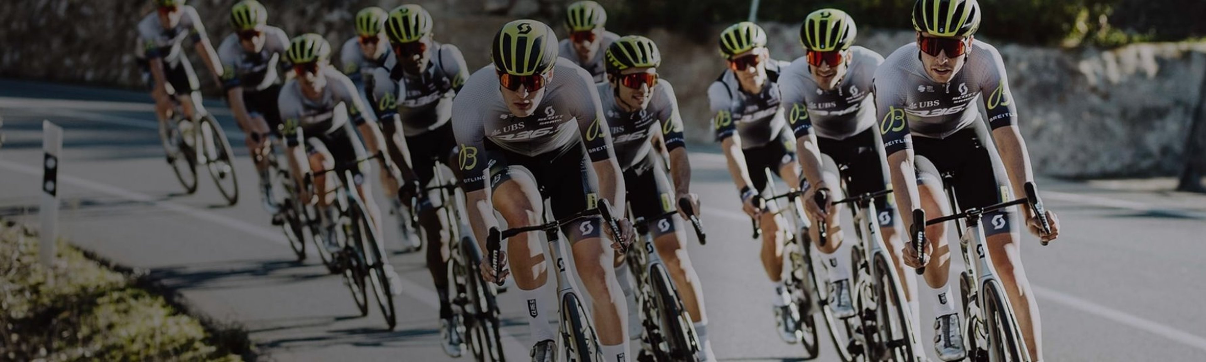 {generated} Cyclists in coordinated team kits with visible Breitling branding ride in formation along a curving sunlit road.