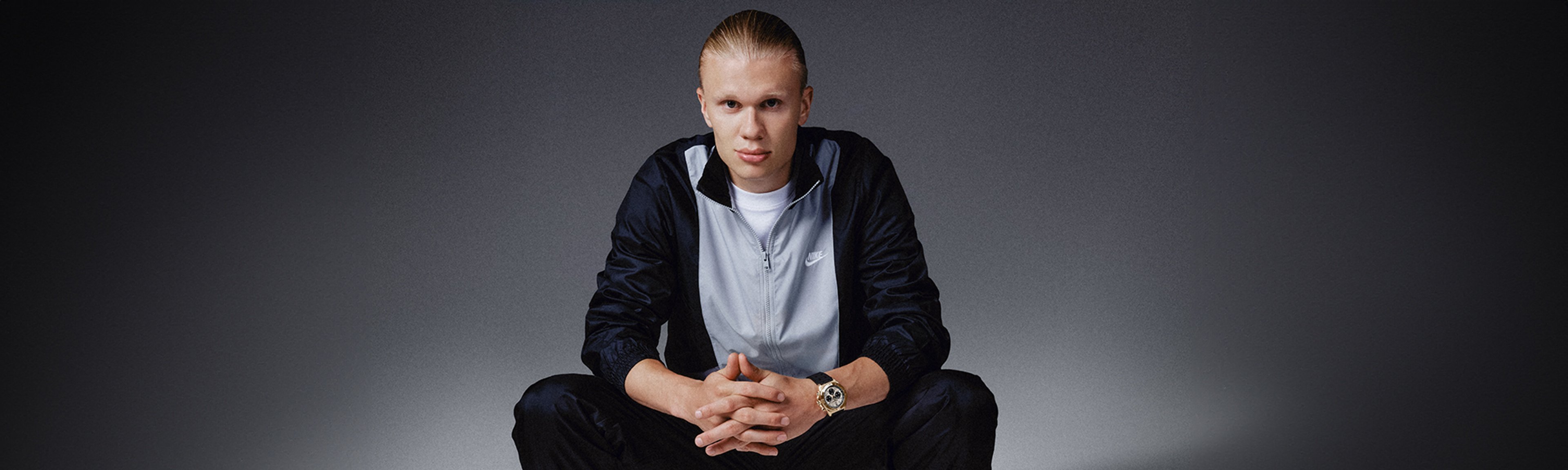 {generated} A seated male ambassador in sportswear highlights a gold Breitling watch against a dark backdrop with a composed posture.