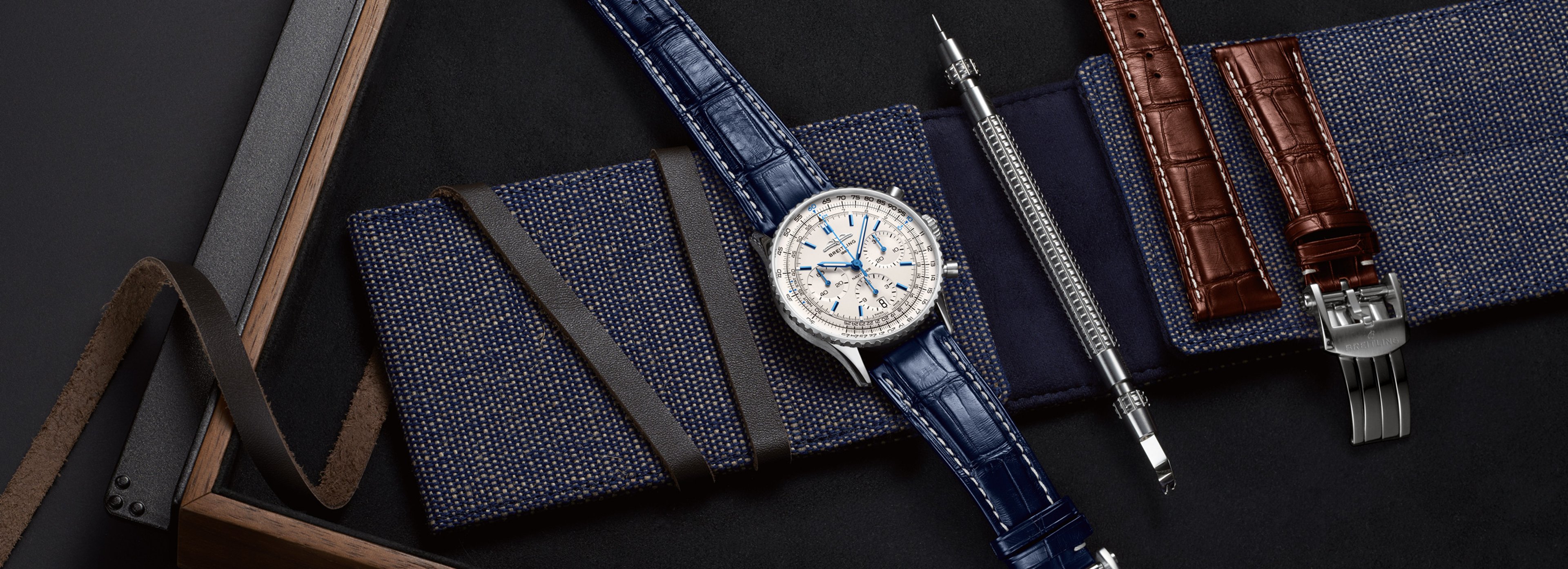 {generated} Breitling watch with blue strap arranged in a Weekender Pack beside spare brown strap and tool on a dark textured surface