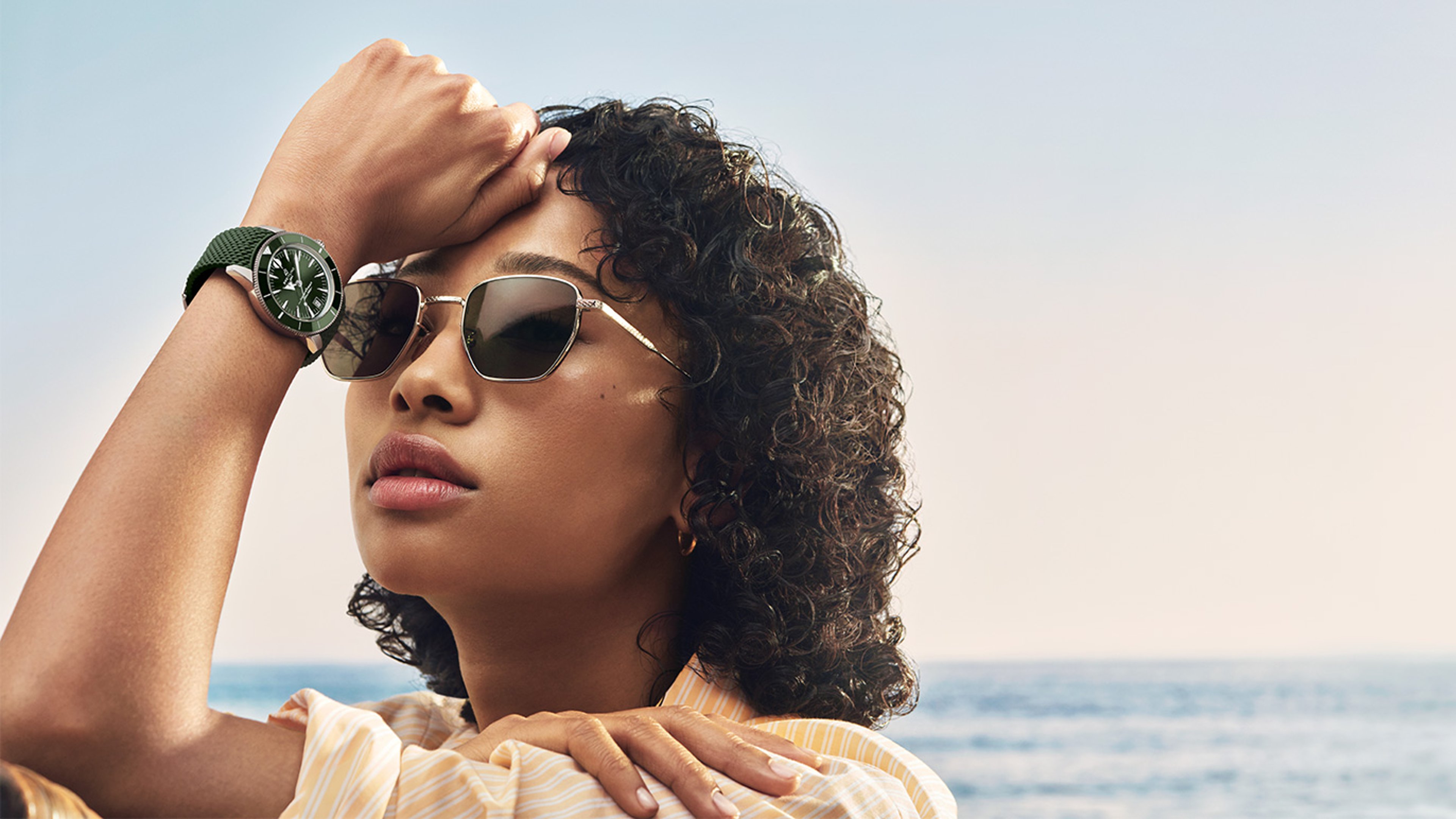 {generated} A person by the sea raises an arm to reveal a Breitling Superocean Heritage watch with a green strap in soft sunlight.