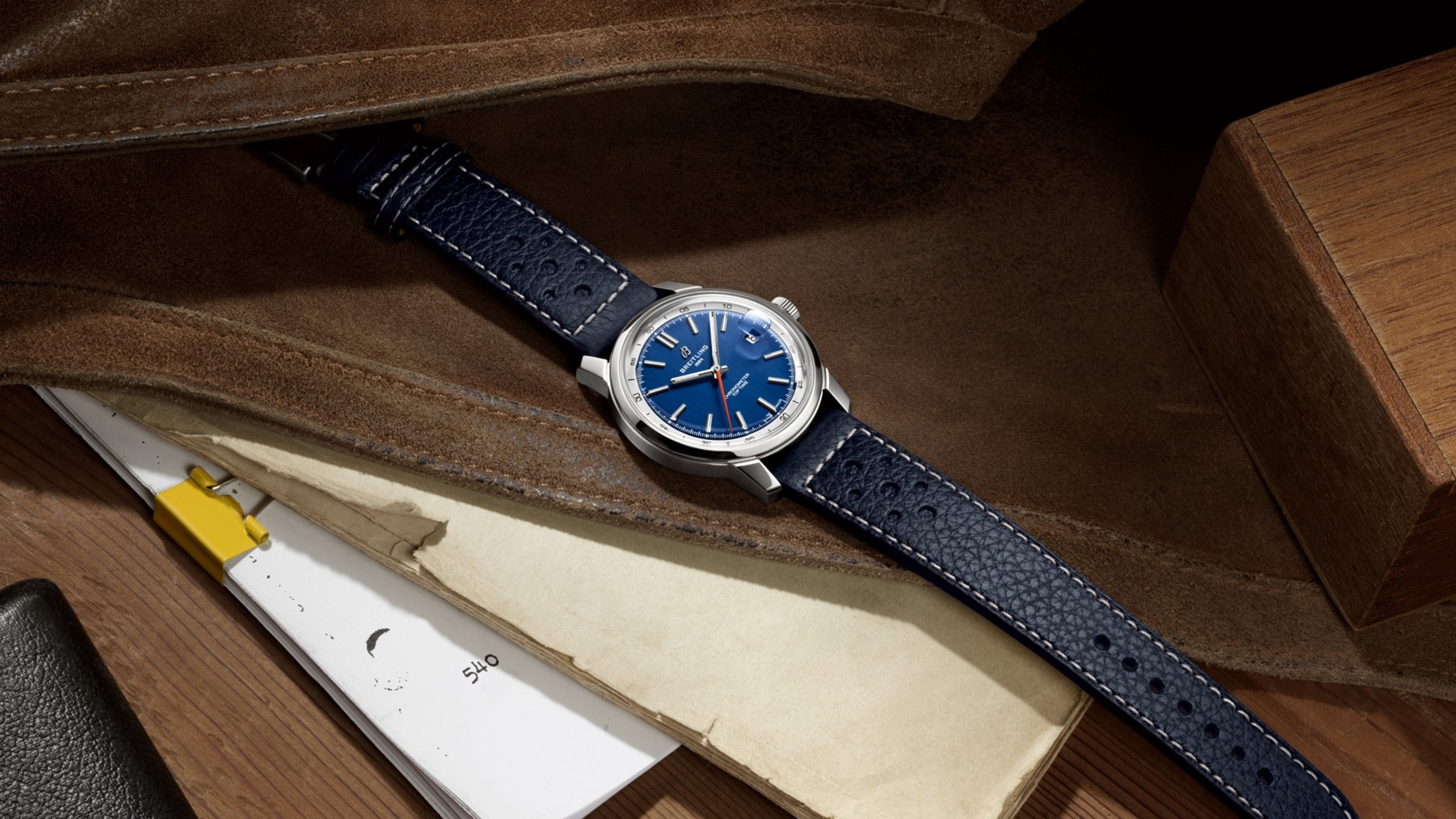 {generated} ブルー文字盤とレザーストラップのBreitling Top Time B31がスエード上に置かれ書類と木製アクセントが添えられる