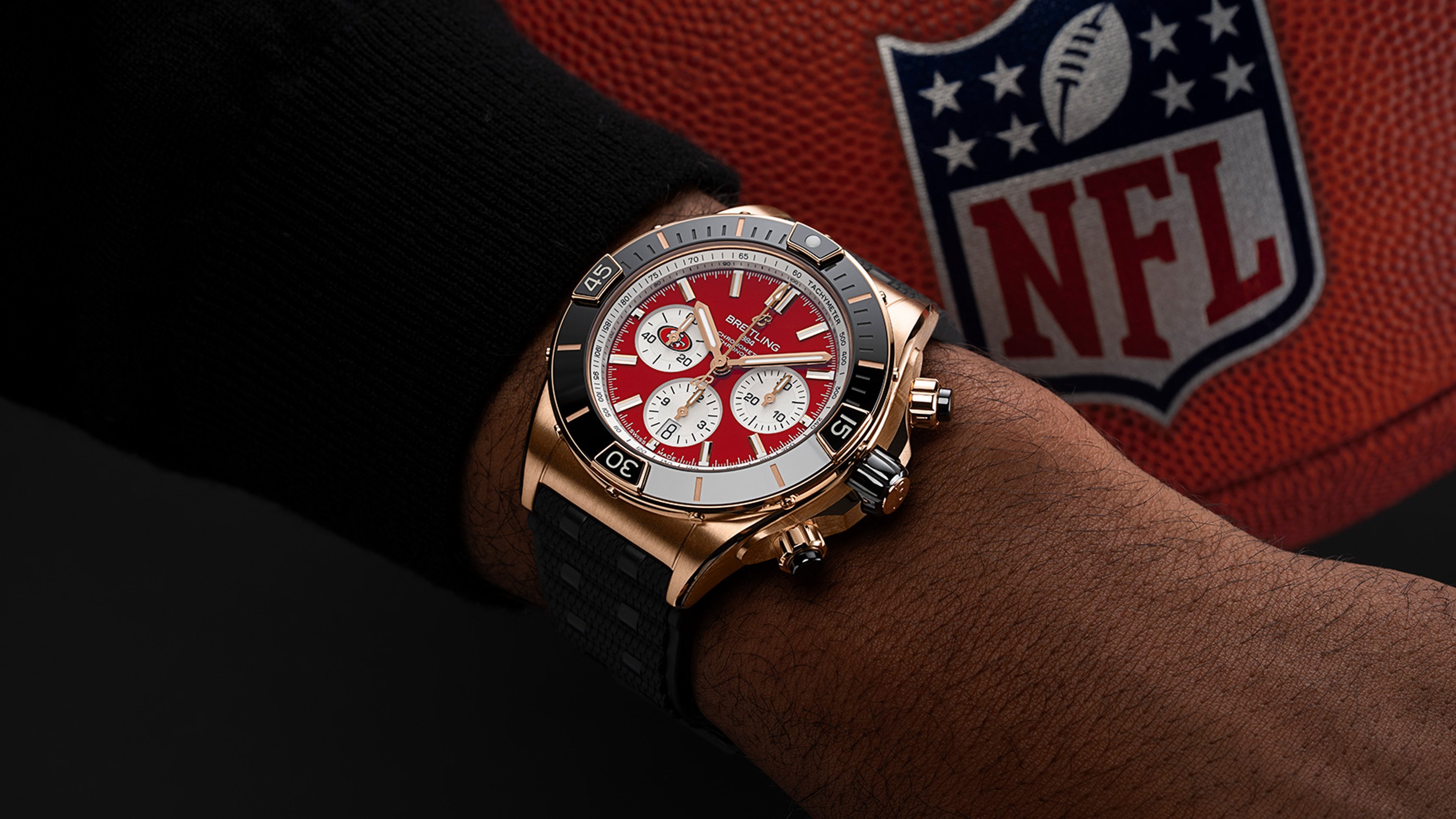 {generated} Cronógrafo Breitling con esfera roja de los San Francisco 49ers en la muñeca, junto a un balón NFL que refuerza el tema del equipo.