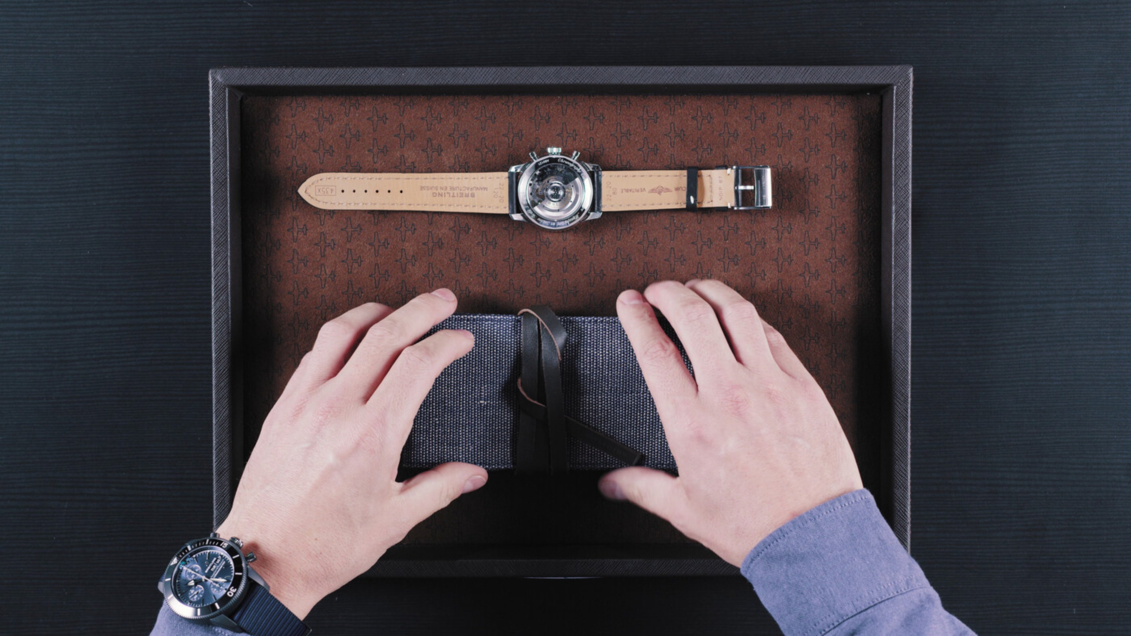 {generated} Hands open a fabric case inside a box with a Breitling watch displayed above, showing its polished caseback and leather strap.