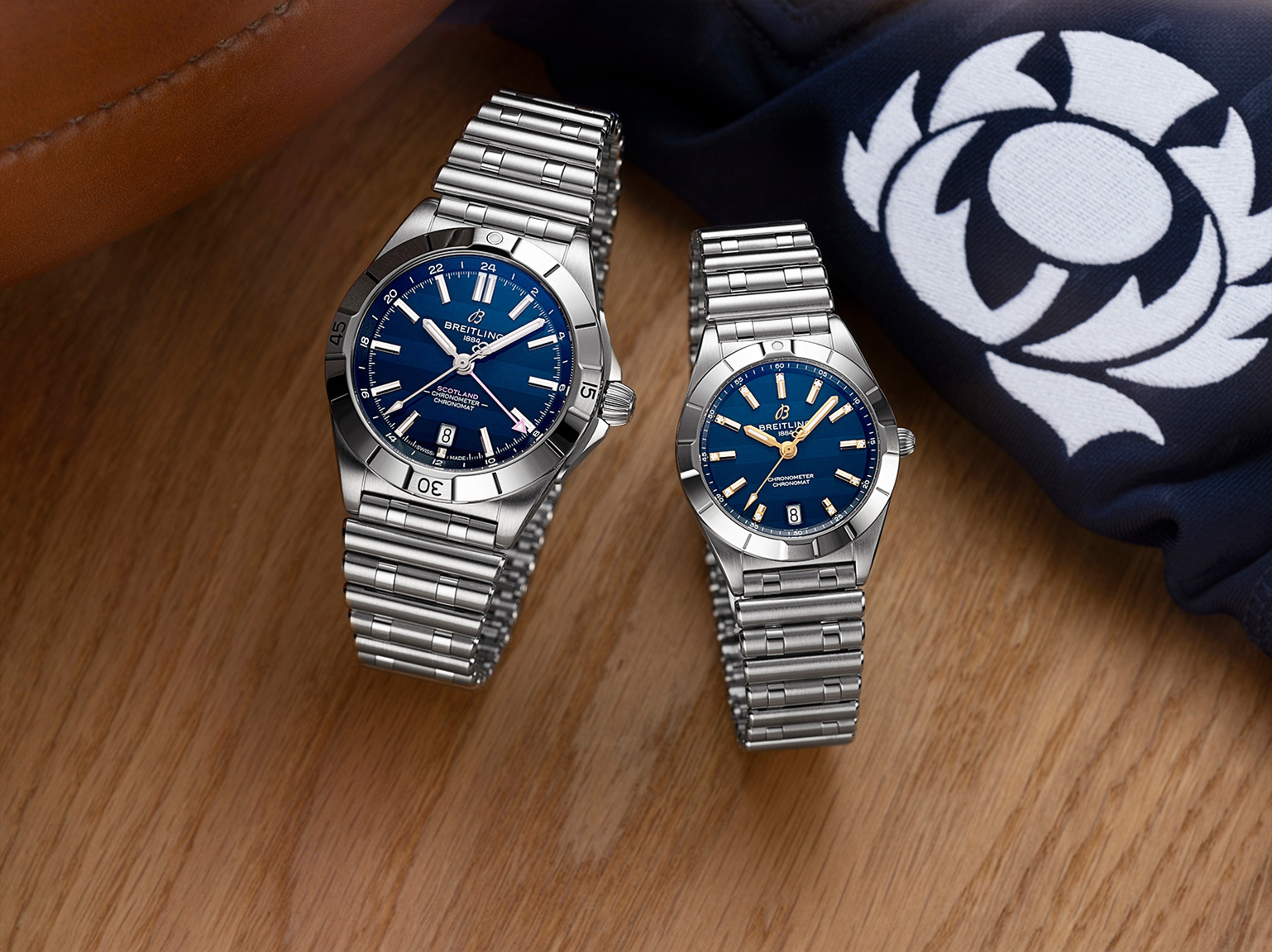 {generated} Two Breitling watches with blue dials and steel bracelets displayed beside a Scotland rugby emblem on a wooden surface.